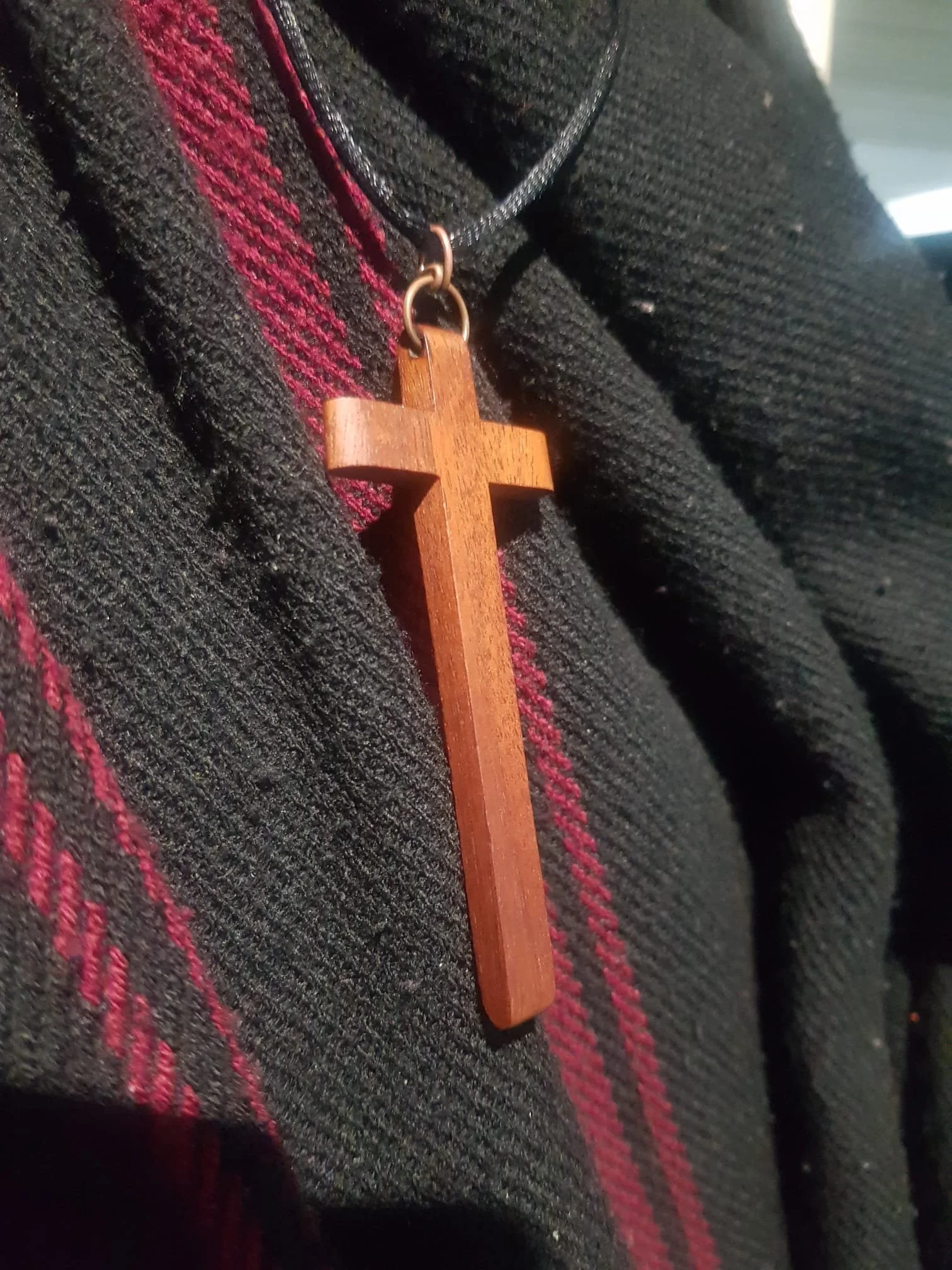 A wooden cross pendant attached to a black cord necklace, resting on a dark, textured fabric with red and black striped pattern.