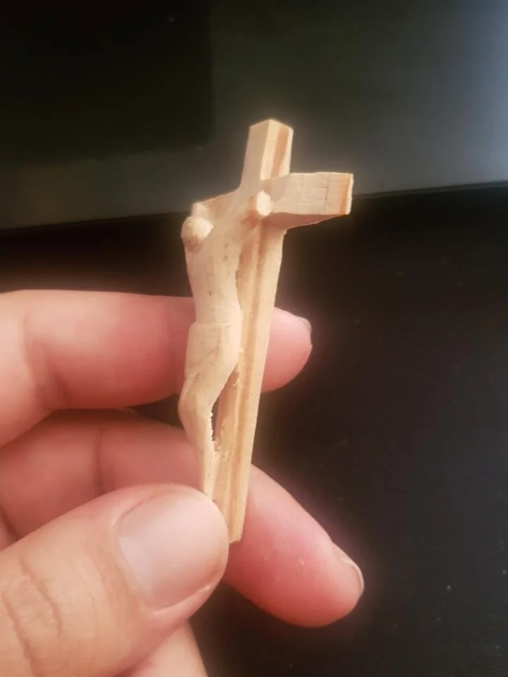 A small wooden figure resembling a person, held between a person's thumb and fingers, with a simple, rough crafted appearance.