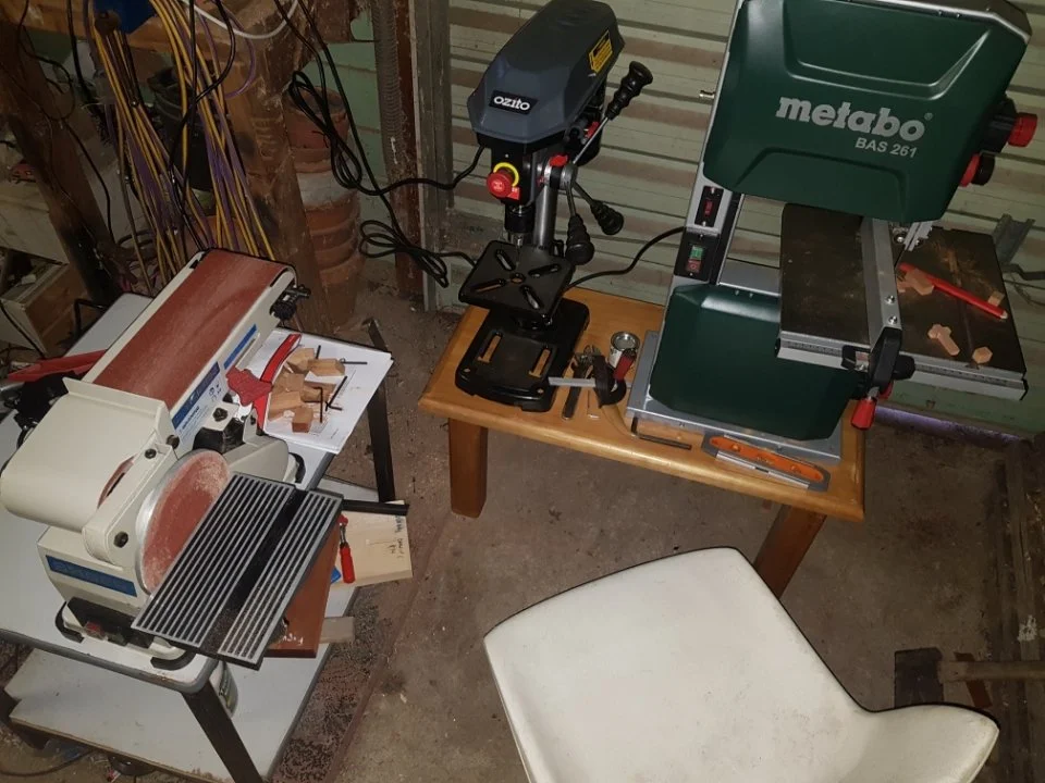 Workshop setup with woodworking tools, including a belt sander, a drillpress, and a small bandsaw on a wooden workbench, with supplies, tools, and a white chair nearby.