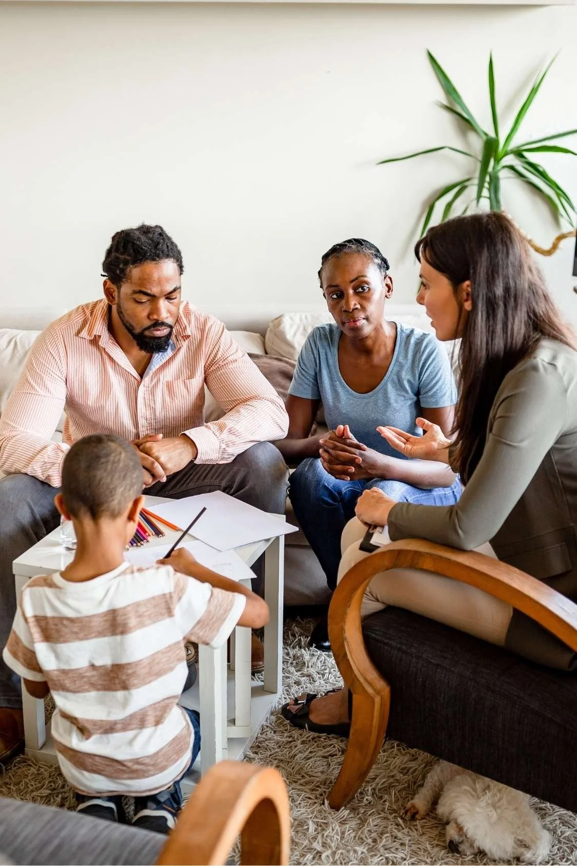 A diverse group of adults and a child having a serious discussion in a living room, with a dog lying on the rug underneath the coffee table.