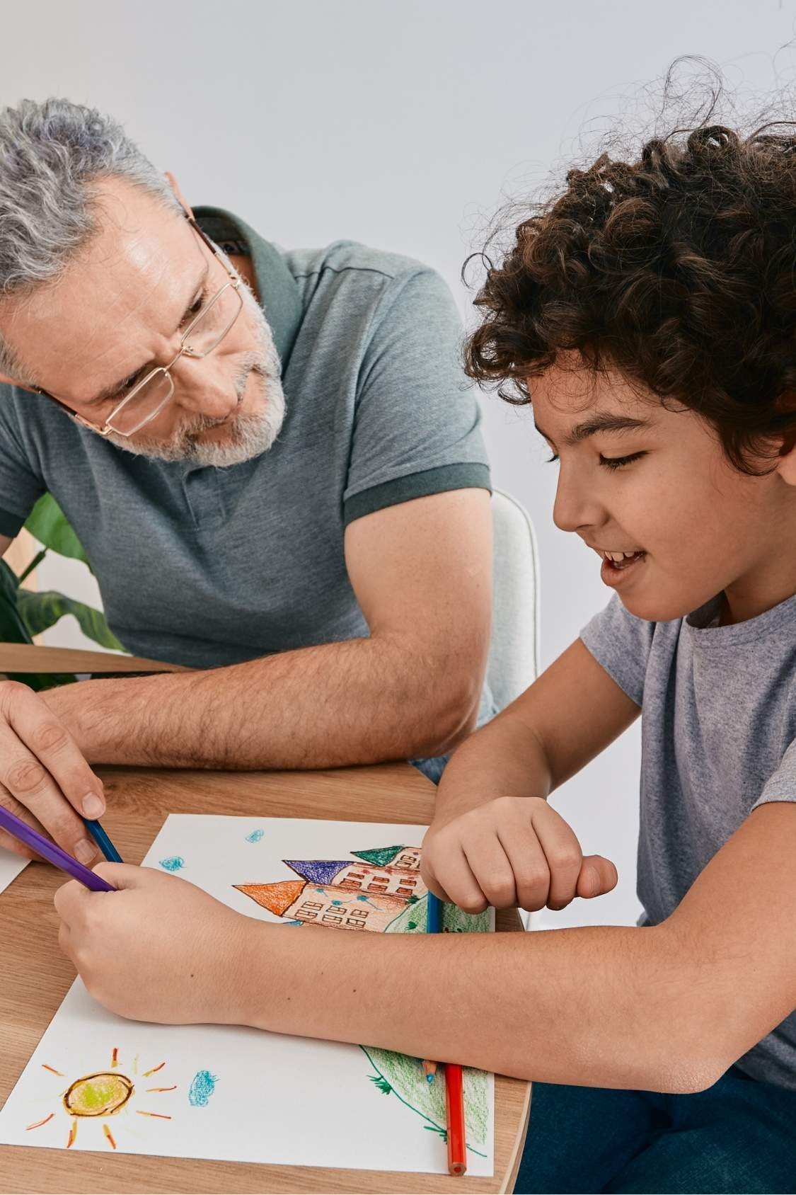 An older man with gray hair and glasses sits at a table, smiling and looking at a young boy who is coloring a picture of a house and trees. The boy has curly brown hair and is focused on his artwork.