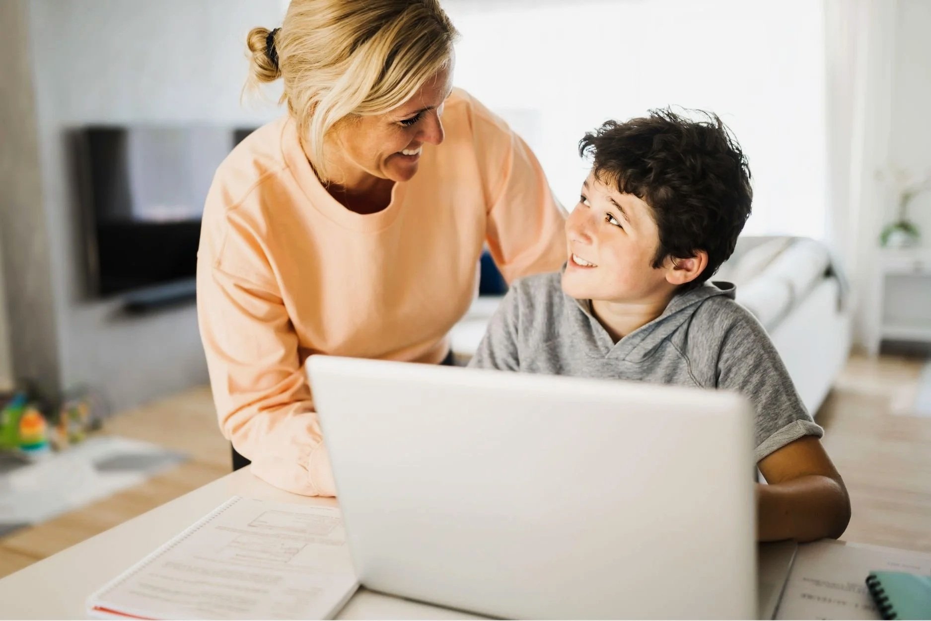 A woman and a young boy smile at each other while sitting at a table with a laptop and papers in front of them, in a bright indoor setting.