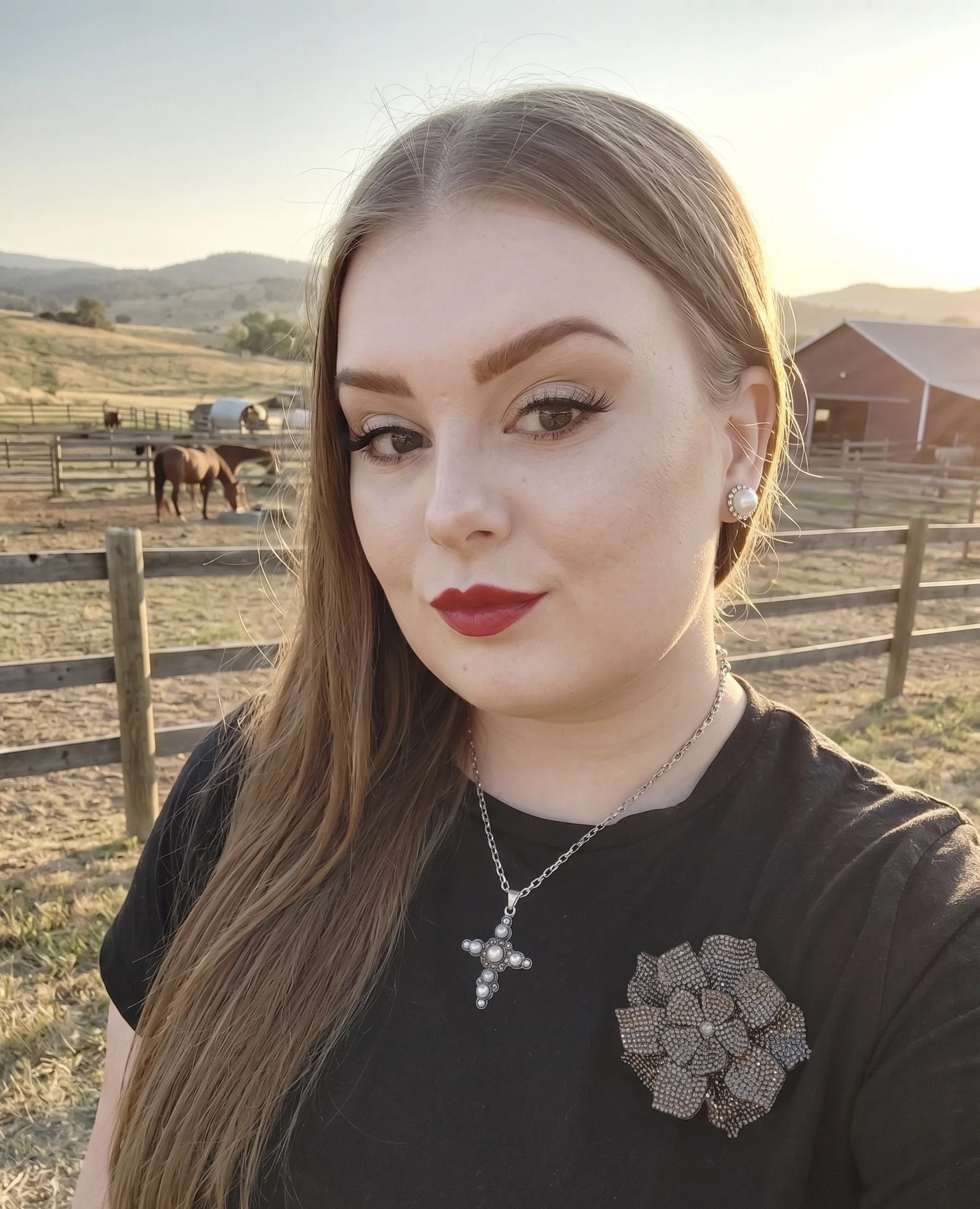 Close-up of a young woman with long hair, wearing makeup, pearl earrings, a statement necklace, and a black t-shirt with a decorative pattern, posing outdoors on a farm with horses and a barn in the background during sunset.