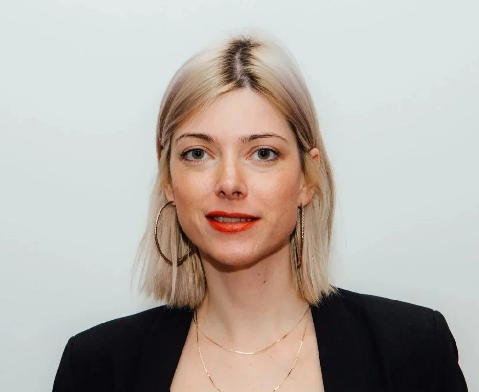 A portrait of a young woman with shoulder-length blonde hair, wearing large hoop earrings, a black blazer, and a delicate gold necklace, standing against a plain light background.