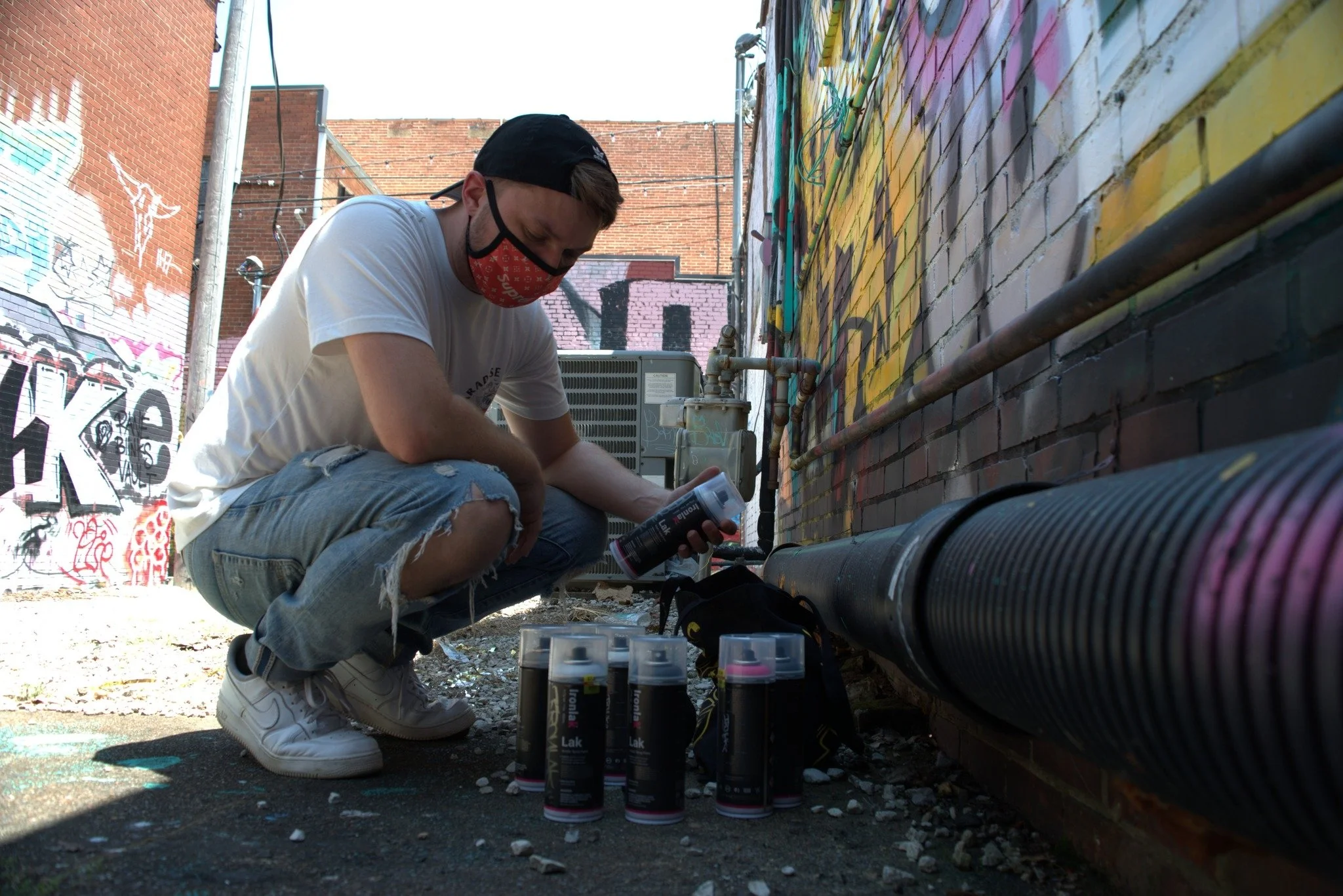 A young man wearing a black baseball cap, red face mask, white T-shirt, and ripped jeans crouches down spray painting graffiti on a brick wall in an alley.