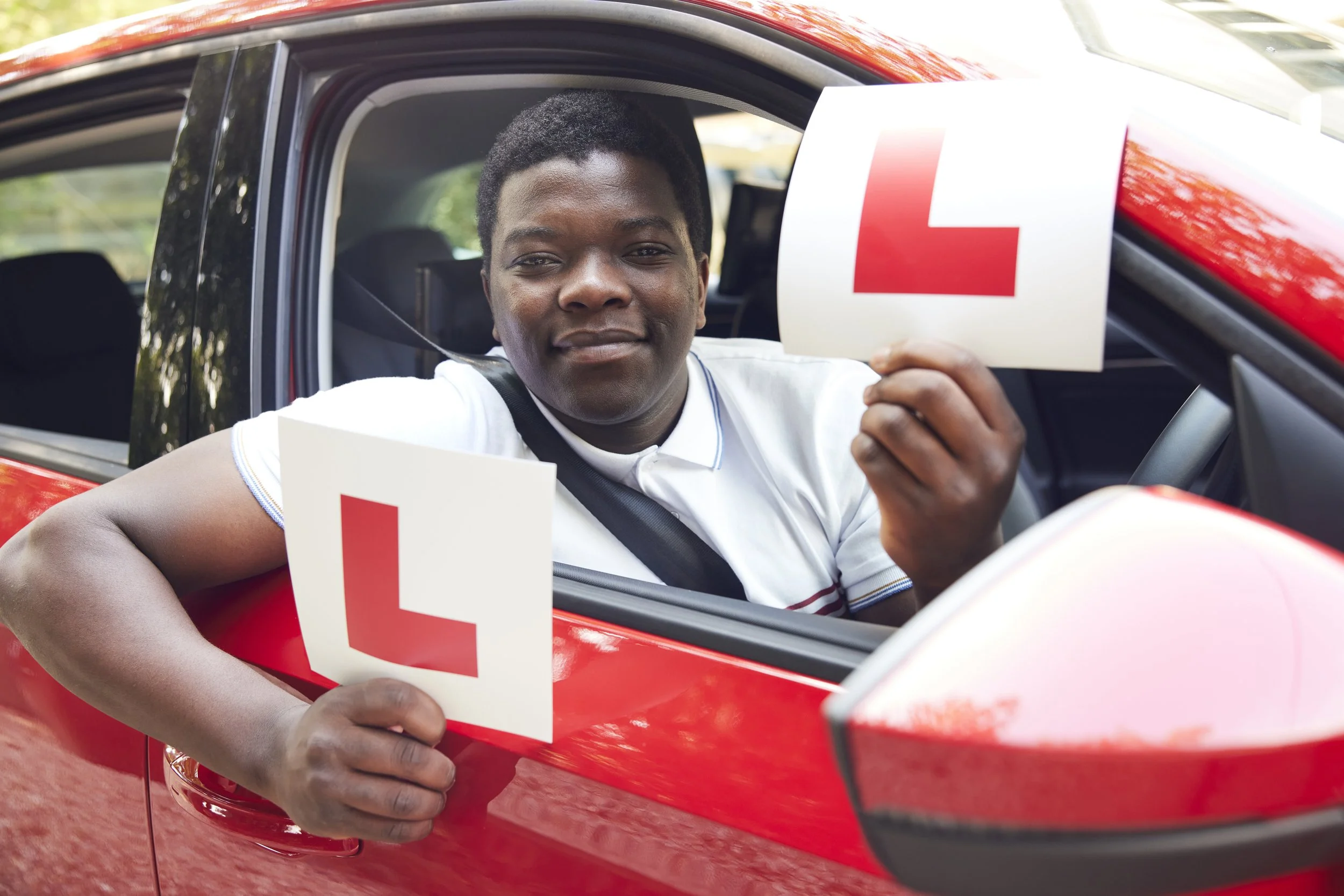 A young man sitting in a red car window, holding a driving exam 'L' sign, with a smile