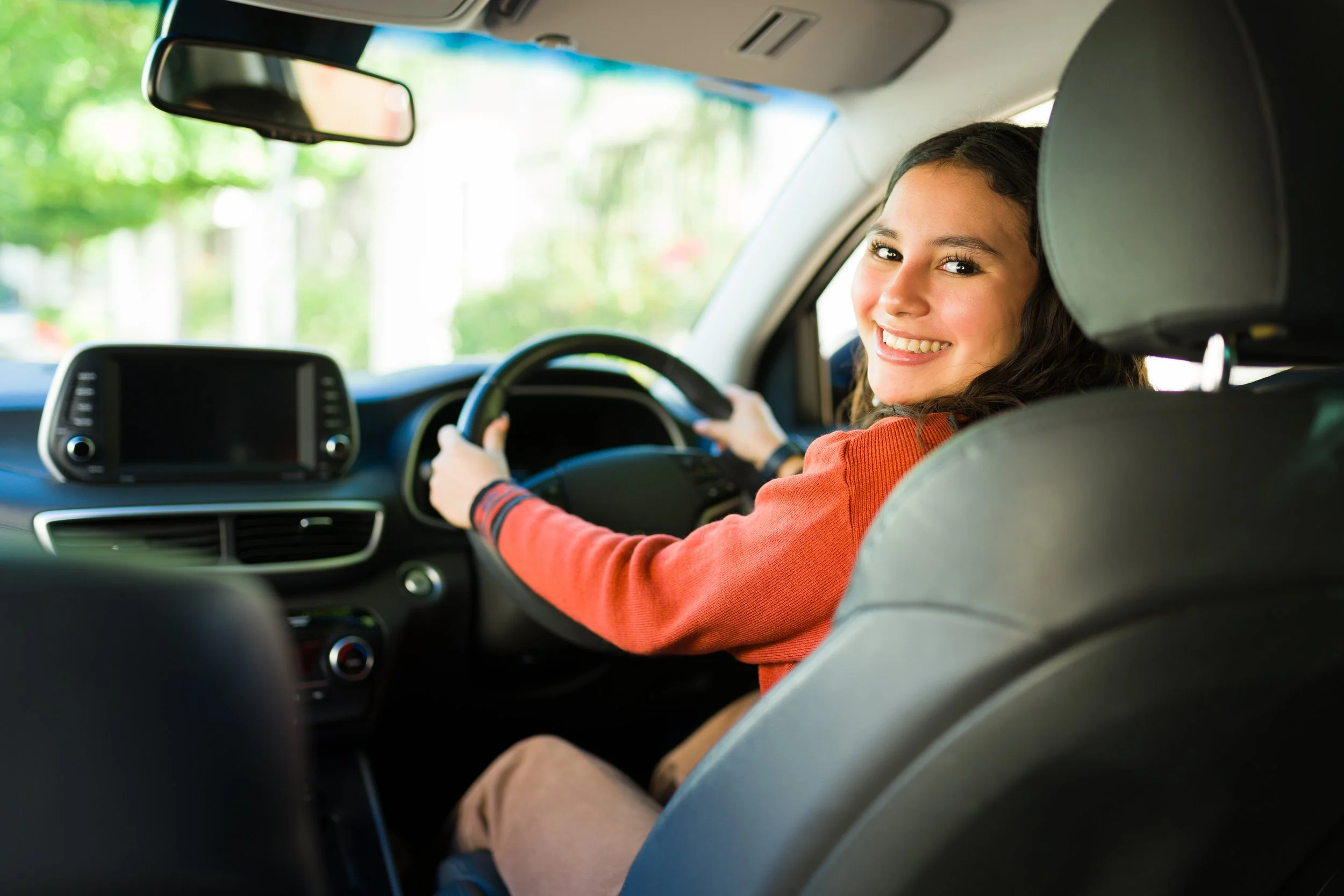 A young woman with long dark hair smiling while driving a car, wearing an orange long-sleeved shirt, with a scenic green outdoor background visible through the windshield.