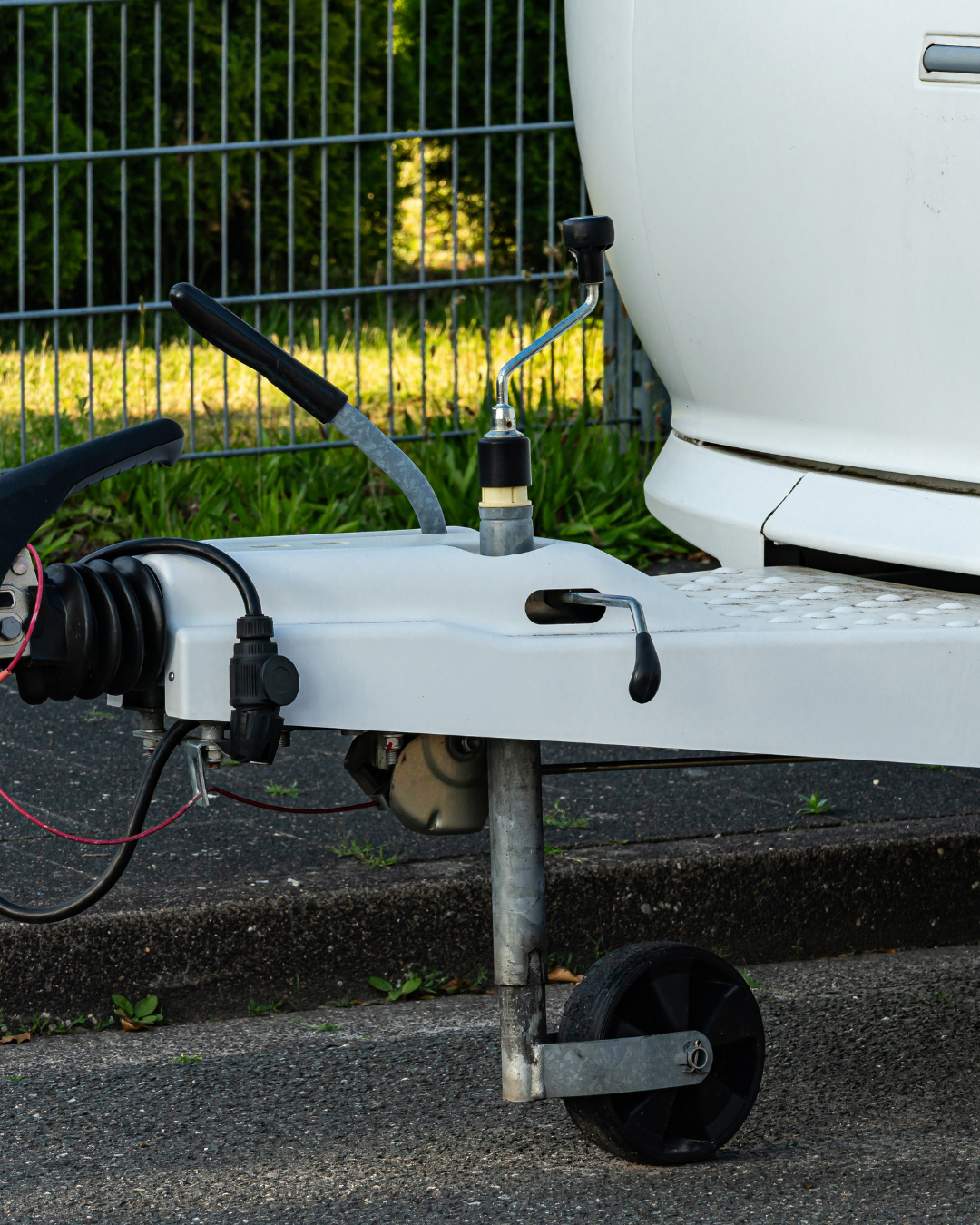 Close-up of a trailer hitch, parked on a paved surface near grass and a fence.
