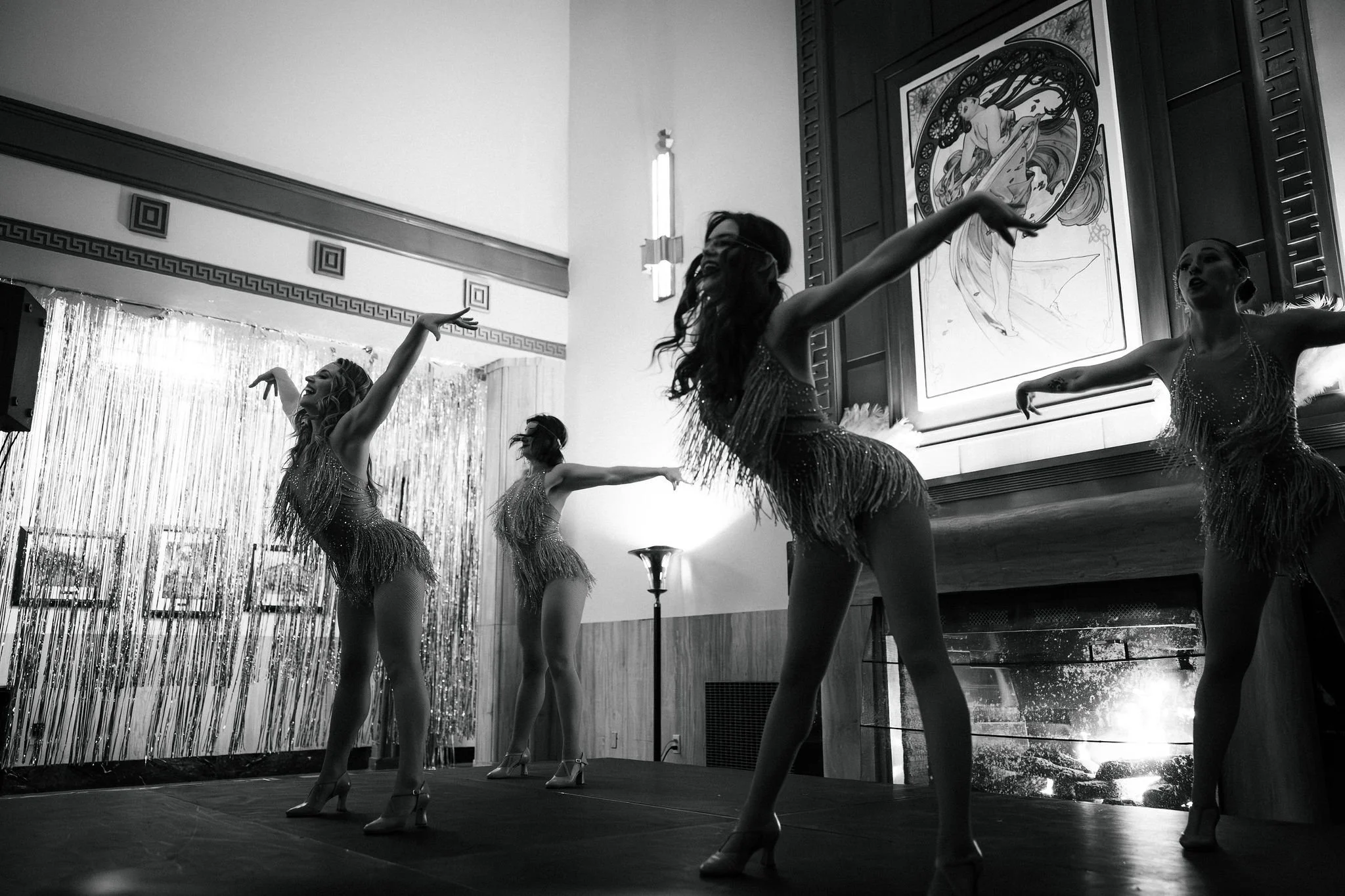 Four dancers in glittery costumes perform on stage indoors, with two large paintings and decorative curtains in the background.