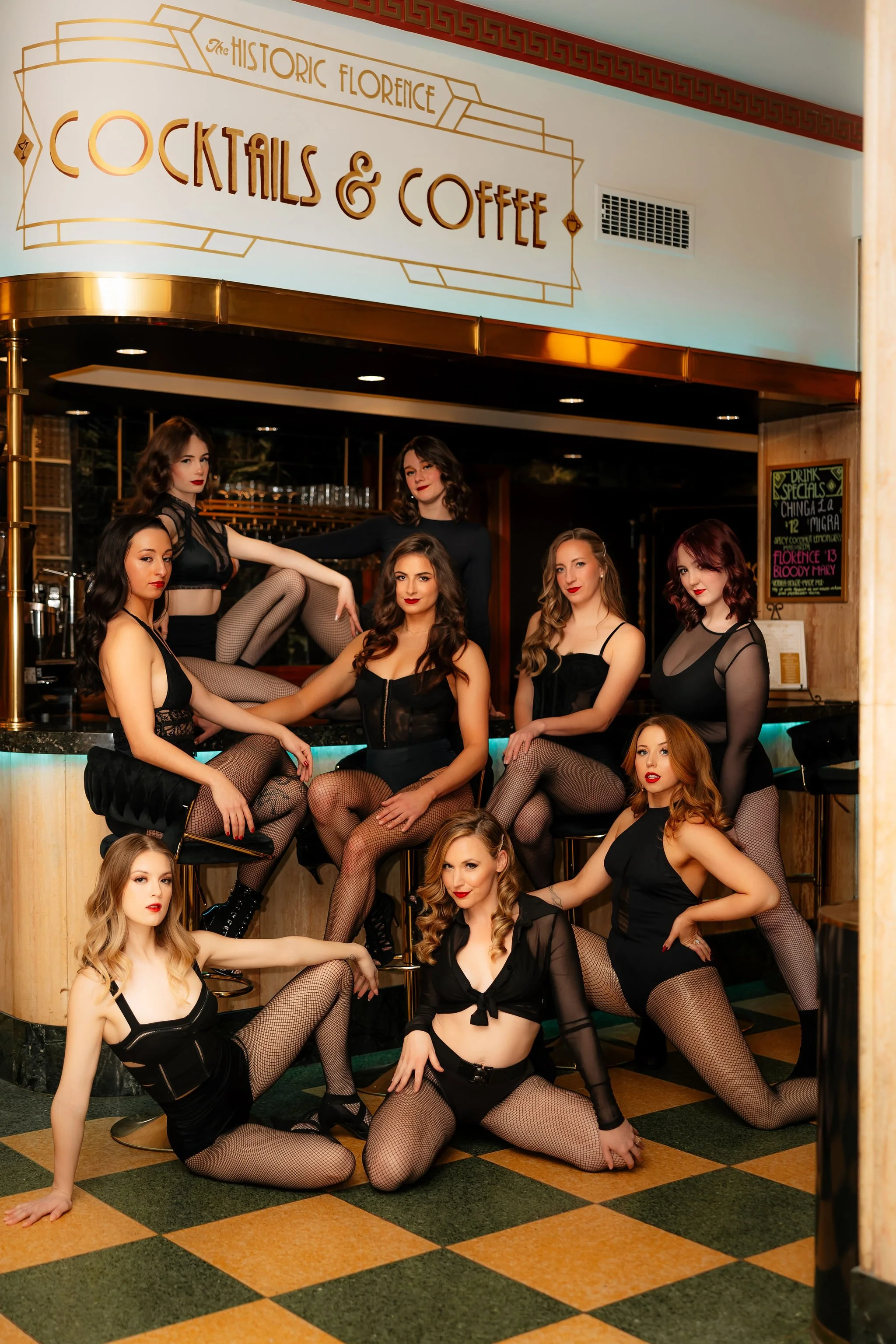 A group of women's fashion models in black lingerie and fishnet stockings posing inside a bar named The Historic Florence Cocktails & Coffee. The bar has a vintage decor with a neon-lit counter, and a chalkboard menu on the wall.