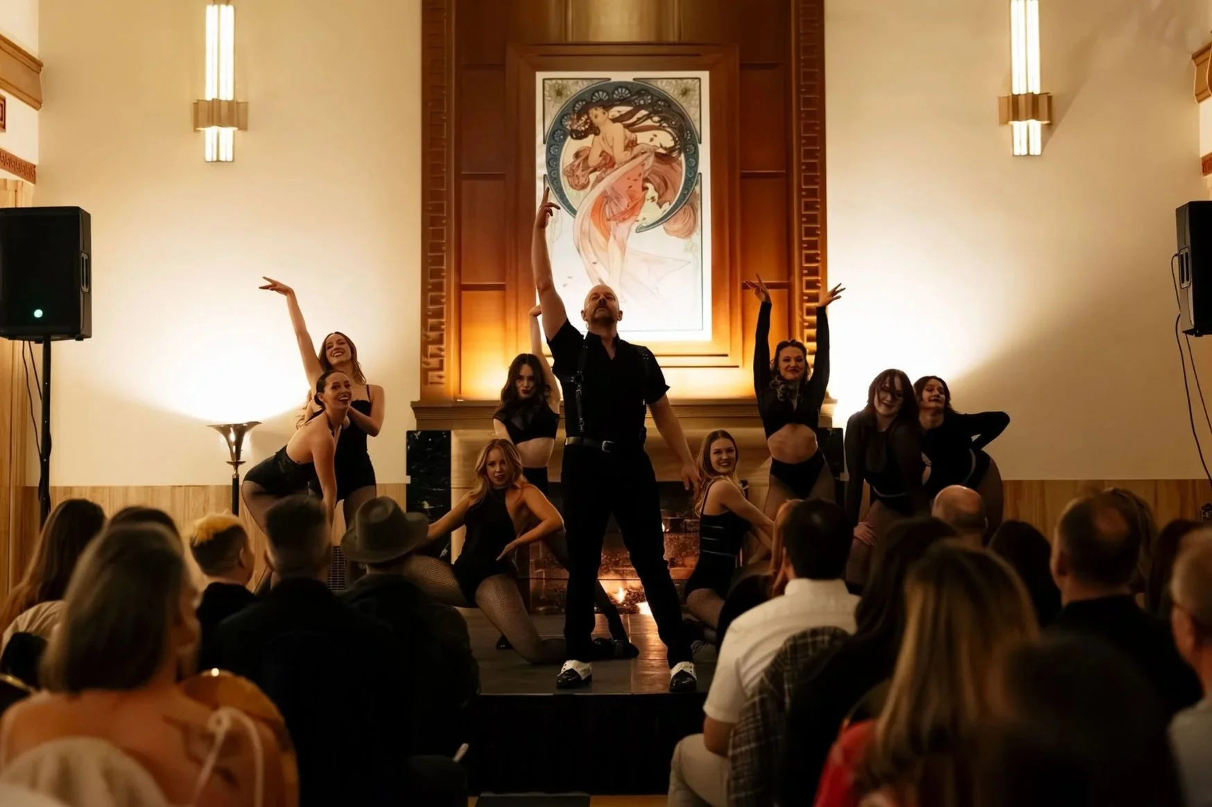 Group of Broadway style performers on stage, with a man in black in the center raising one hand, surrounded by women in black outfits, in front of an audience, at the Historic Florence in Missoula, MT.
