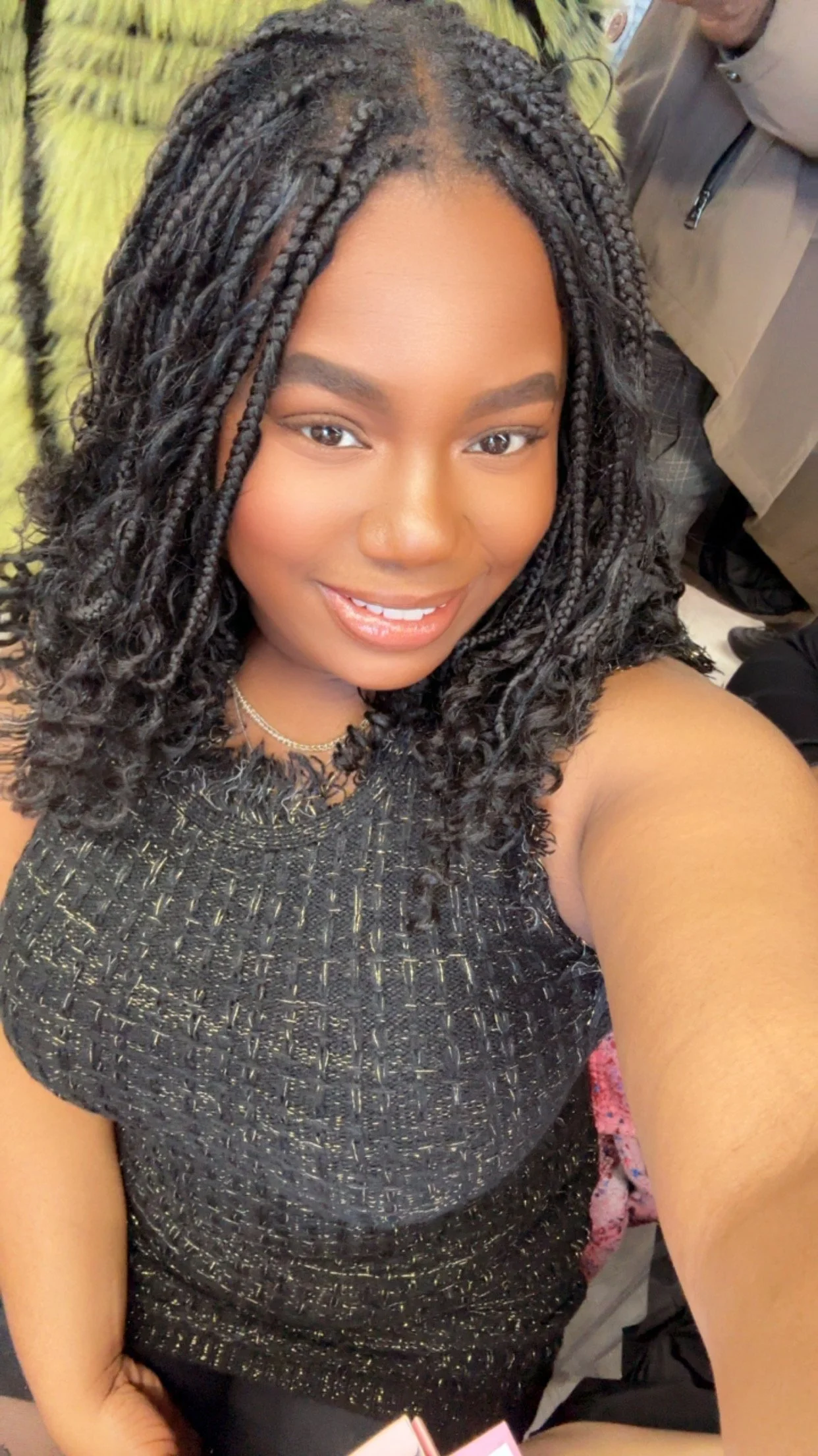 A young woman with medium skin and curly braided hair taking a selfie. She is smiling and wearing a black and gold dress.