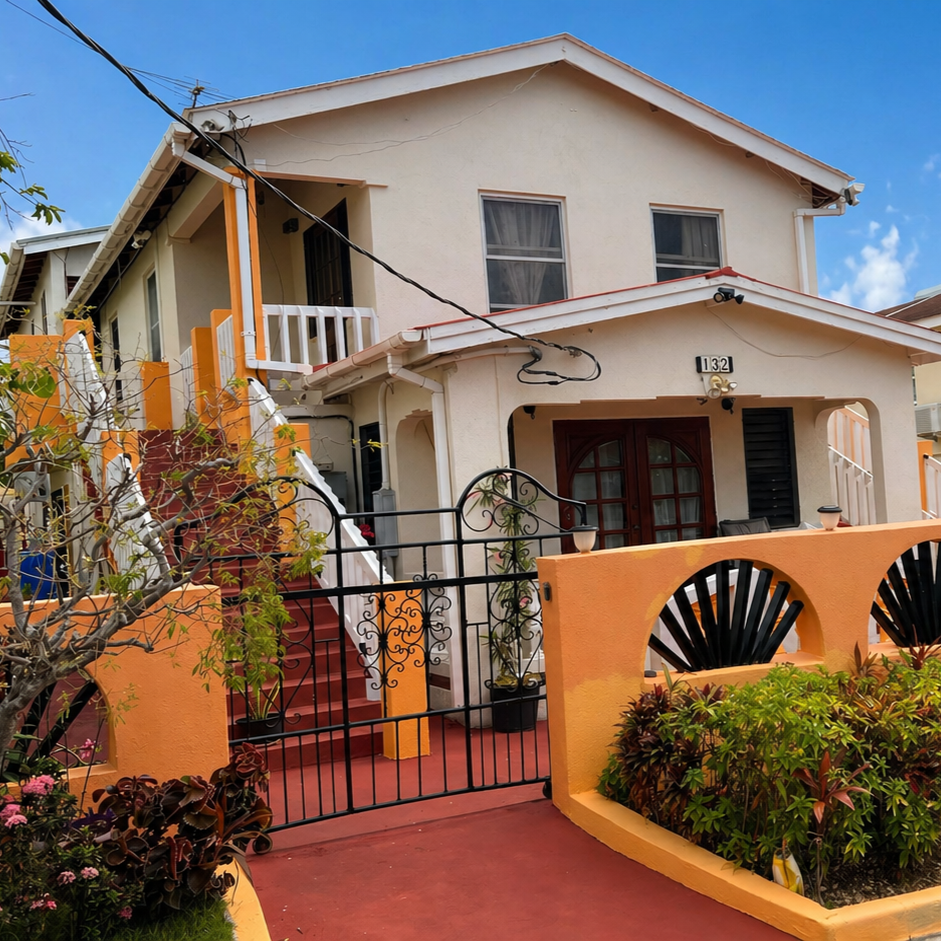 Le Marida Suites - A two-storey house with a cream exterior, brown front door, and red stairs. The house has a small balcony, black window shutters, and a decorative orange and black fence and wall with plants and flowers in the yard.