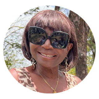 Close-up of your Le Marida Suites Host, Maria, with short brown hair wearing large sunglasses, earrings, and a necklace, smiling outdoors against a background of trees.
