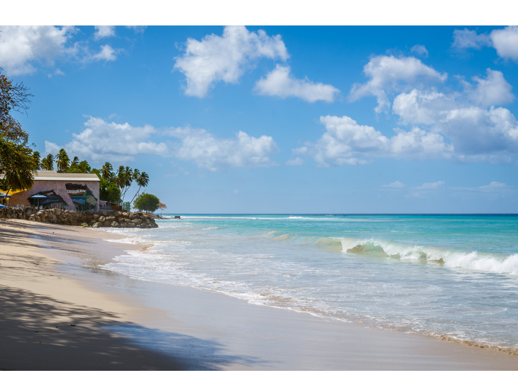 Tropical beach with white sand, clear turquoise water, palm trees, a building with colorful umbrellas, and a blue sky with scattered clouds.