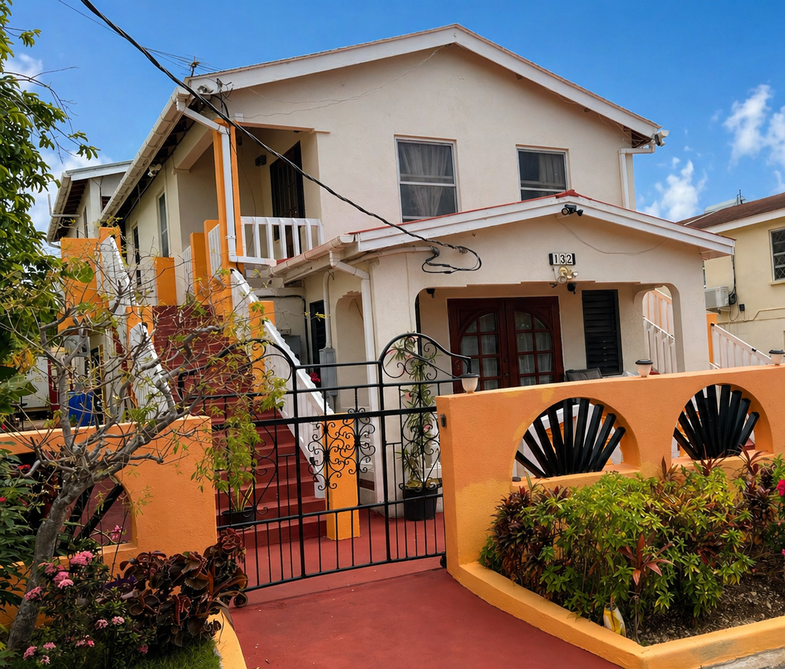 Colorful two-story house with a small front yard enclosed by a fence, red stairs leading to the front door, and plants and bushes in the yard. Blue sky with a few clouds above.