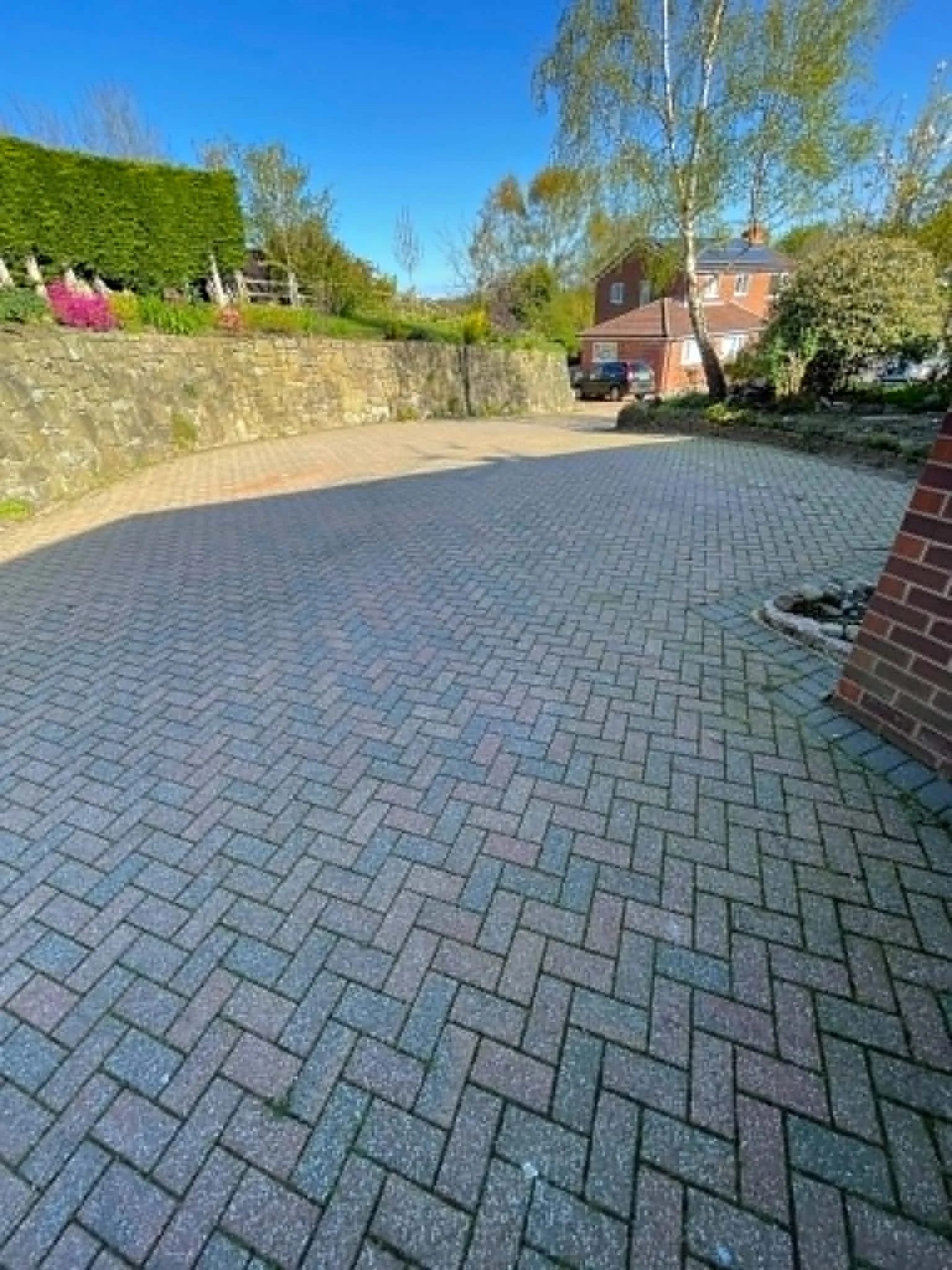 A paved driveway with brick and stone pavers, bordered by a brick wall and a garden with trees and bushes, leading to residential houses under a blue sky.