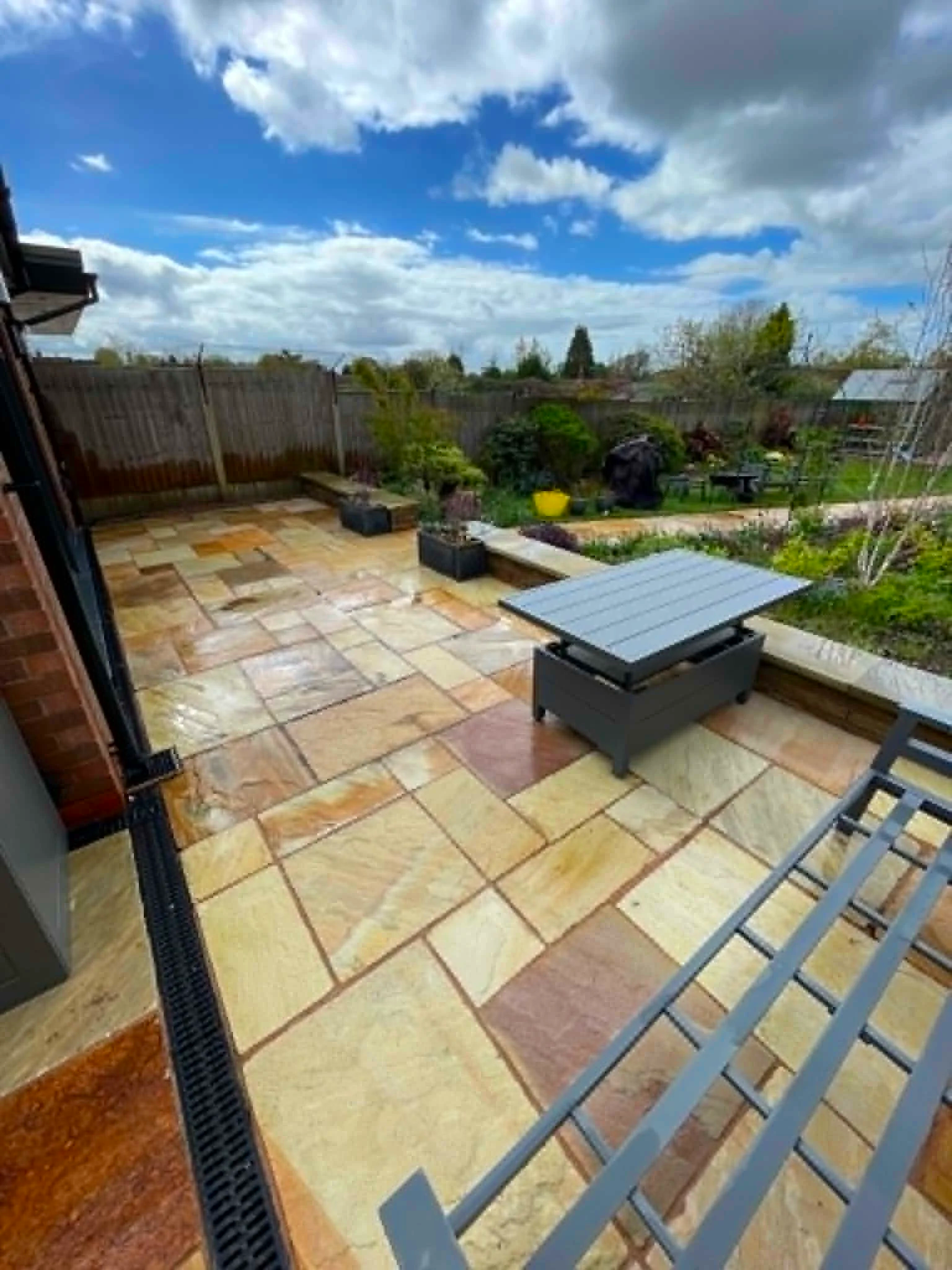 Backyard patio with stone floor, wooden fence, green bushes, and chairs, under partly cloudy sky.