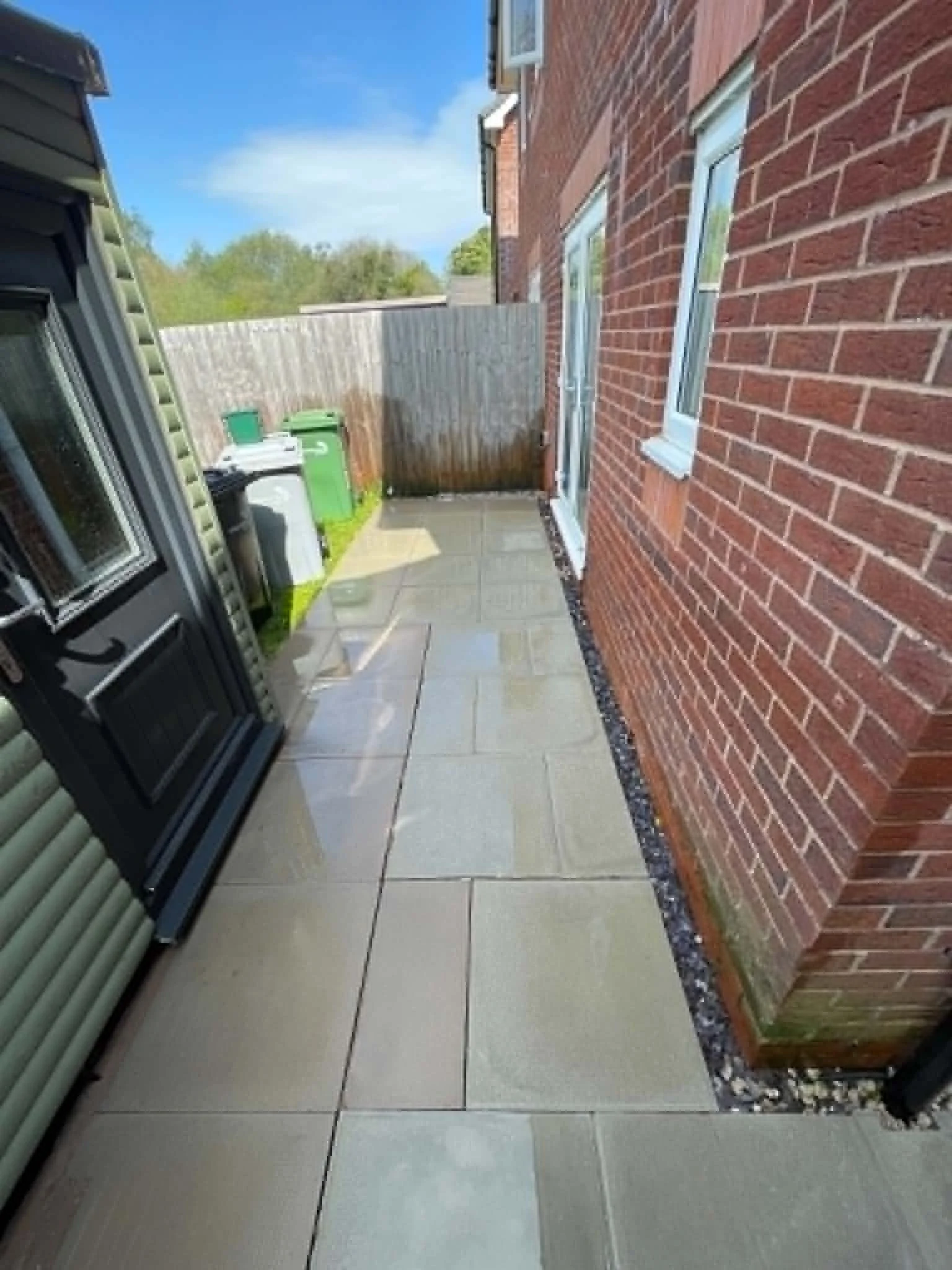 A small backyard patio area with beige tiles, bordered by a red brick wall on the right side and a wooden fence at the back, with some greenery and trees visible beyond the fence. There's a black barbecue grill on the left, next to a green trash bin and a small grey container, under a window on the brick wall. The sky is blue with some clouds.