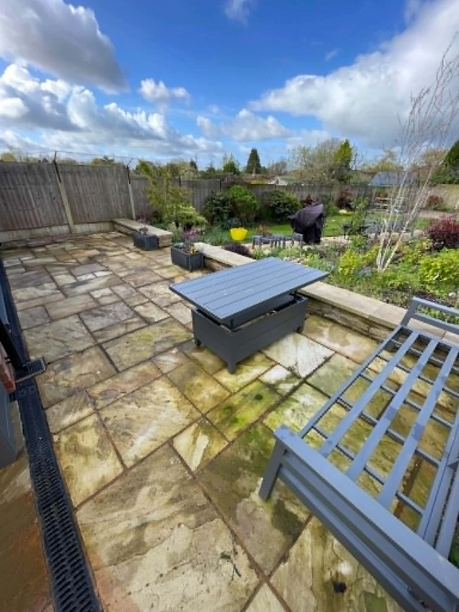 View of a backyard patio with stone flooring, garden beds, and wooden fence, under a partly cloudy sky.