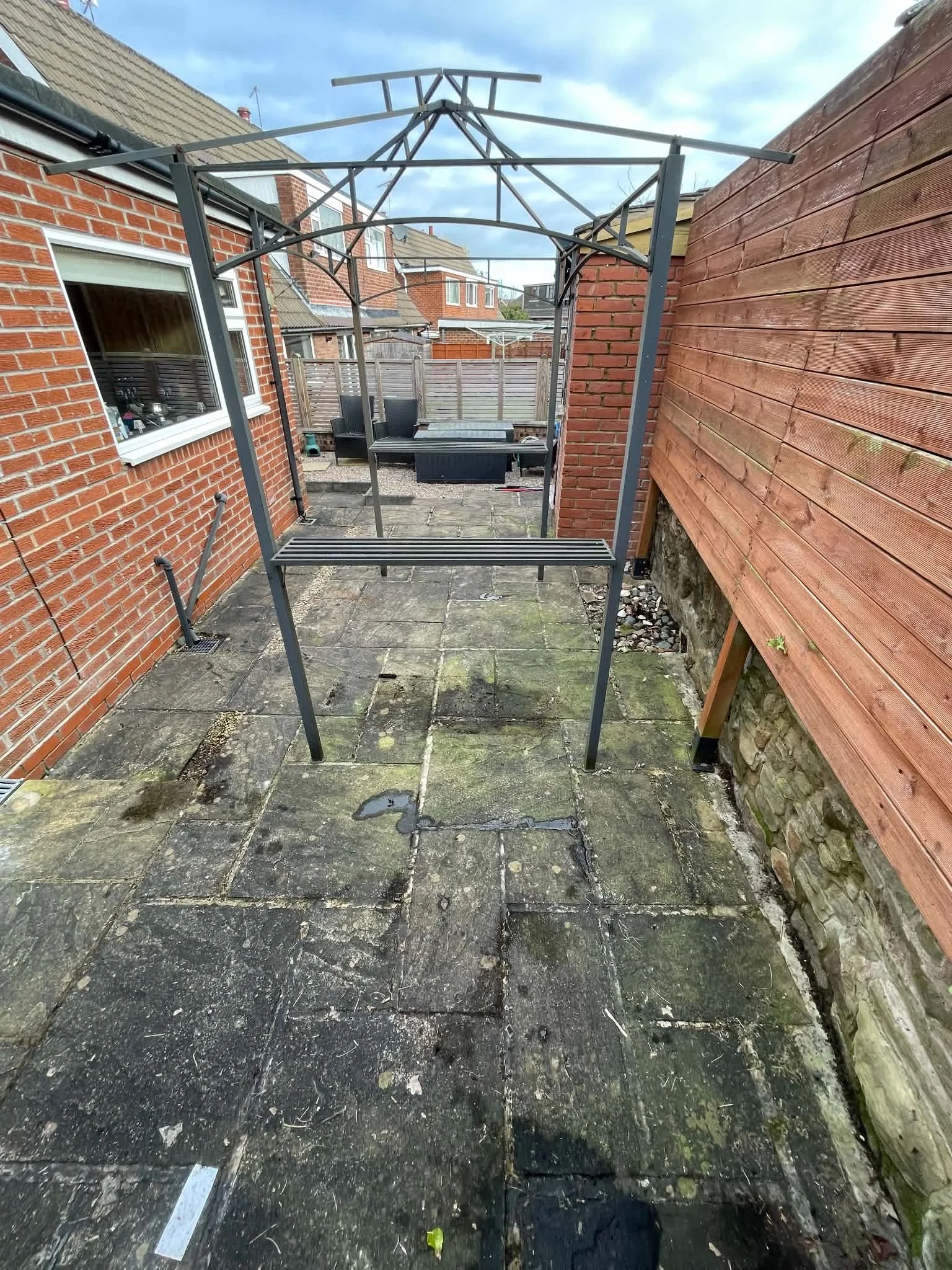 A small outdoor patio area with faded, weathered concrete paving stones, a black metal carport frame without a cover, old brick walls on the left and right sides, and a wooden privacy fence at the back. There is outdoor furniture visible in the background, along with a cloudy sky overhead.