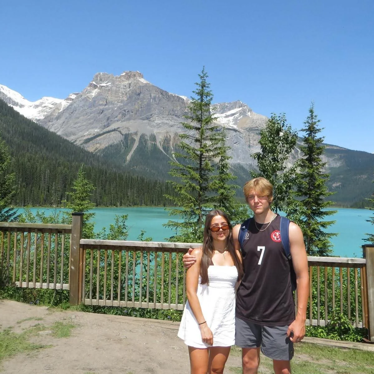 A couple standing in front of a scenic mountain lake with snow-capped peaks, green pine trees, and a wooden fence. The woman is wearing a white dress and sunglasses, and the man is wearing a black sports jersey and gray shorts.