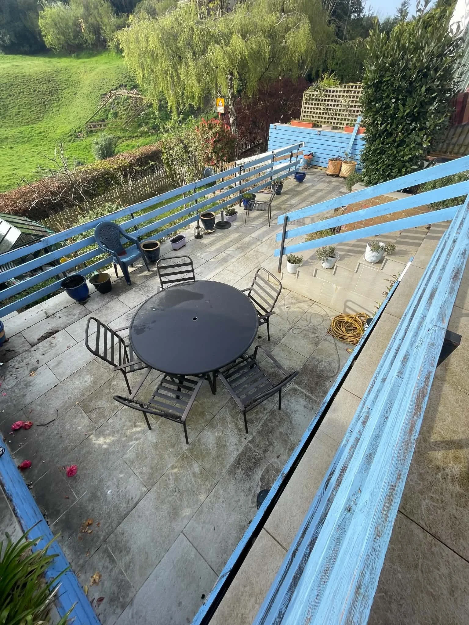 A backyard patio with a round black table and black chairs, surrounded by potted plants and a wooden railing painted blue. The yard is lush green with trees and bushes, and a wooden staircase is visible leading into the yard.
