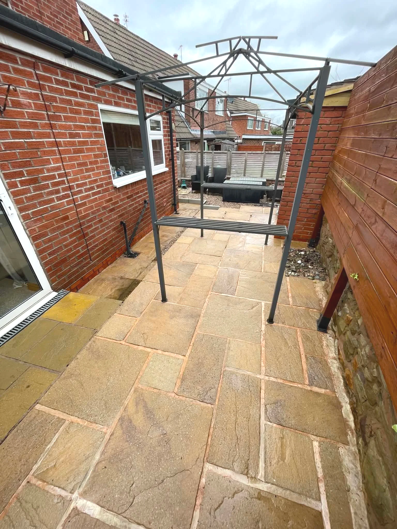 Empty backyard patio with paving stones, a metal gazebo frame, brick house wall on the left, wooden fence on the right, and outdoor seating in the background.