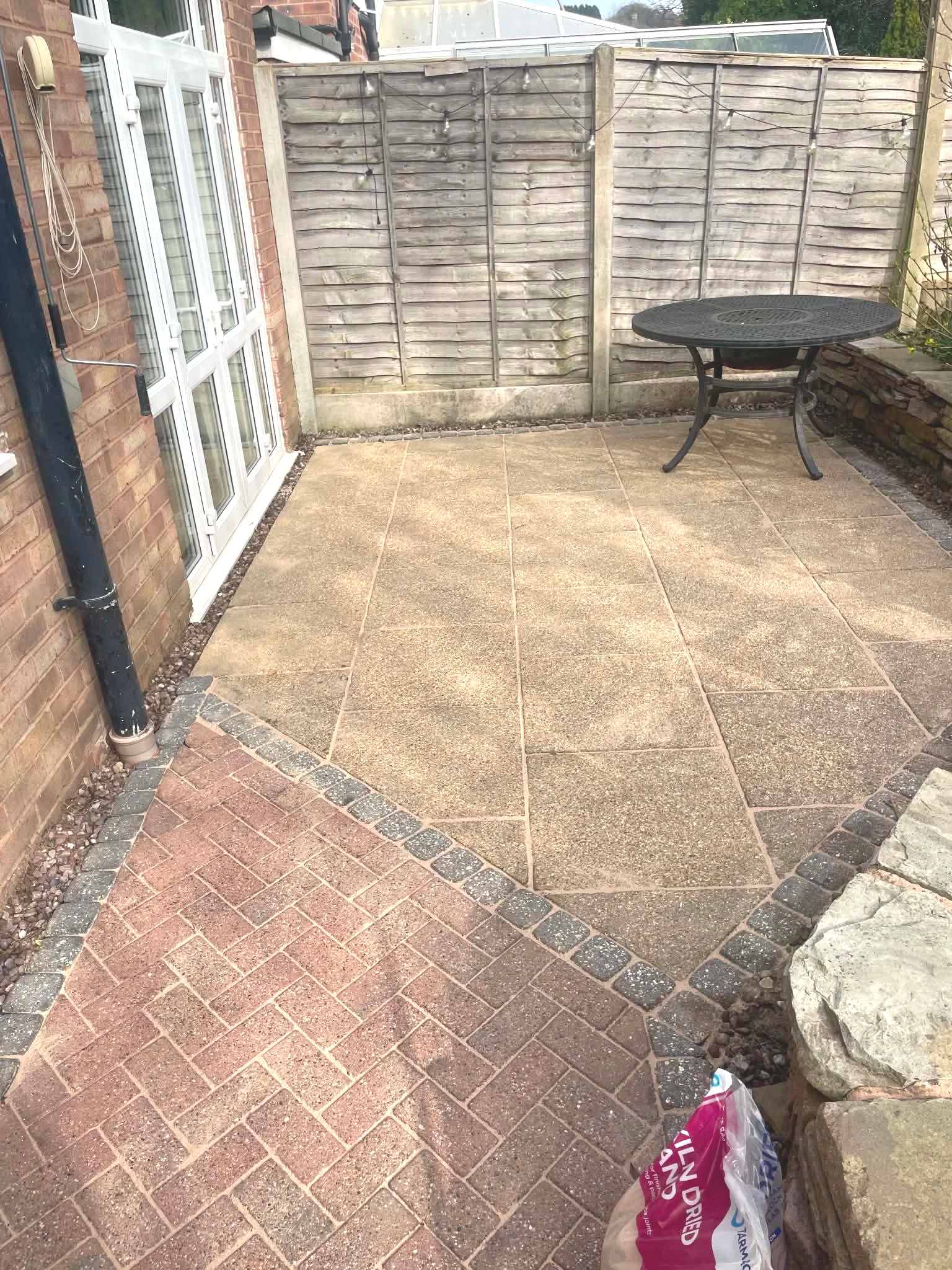 A small outdoor patio or backyard area with a mix of brick and wooden fencing. The ground is covered with square outdoor tiles and a path of smaller brick pavers. There is a black metal table with decorative legs near the wooden fence on the right side. A plastic shopping bag is on the bottom right corner of the image. The left side of the image features a white-framed glass door with a black vertical pipe running down the brick wall next to it.
