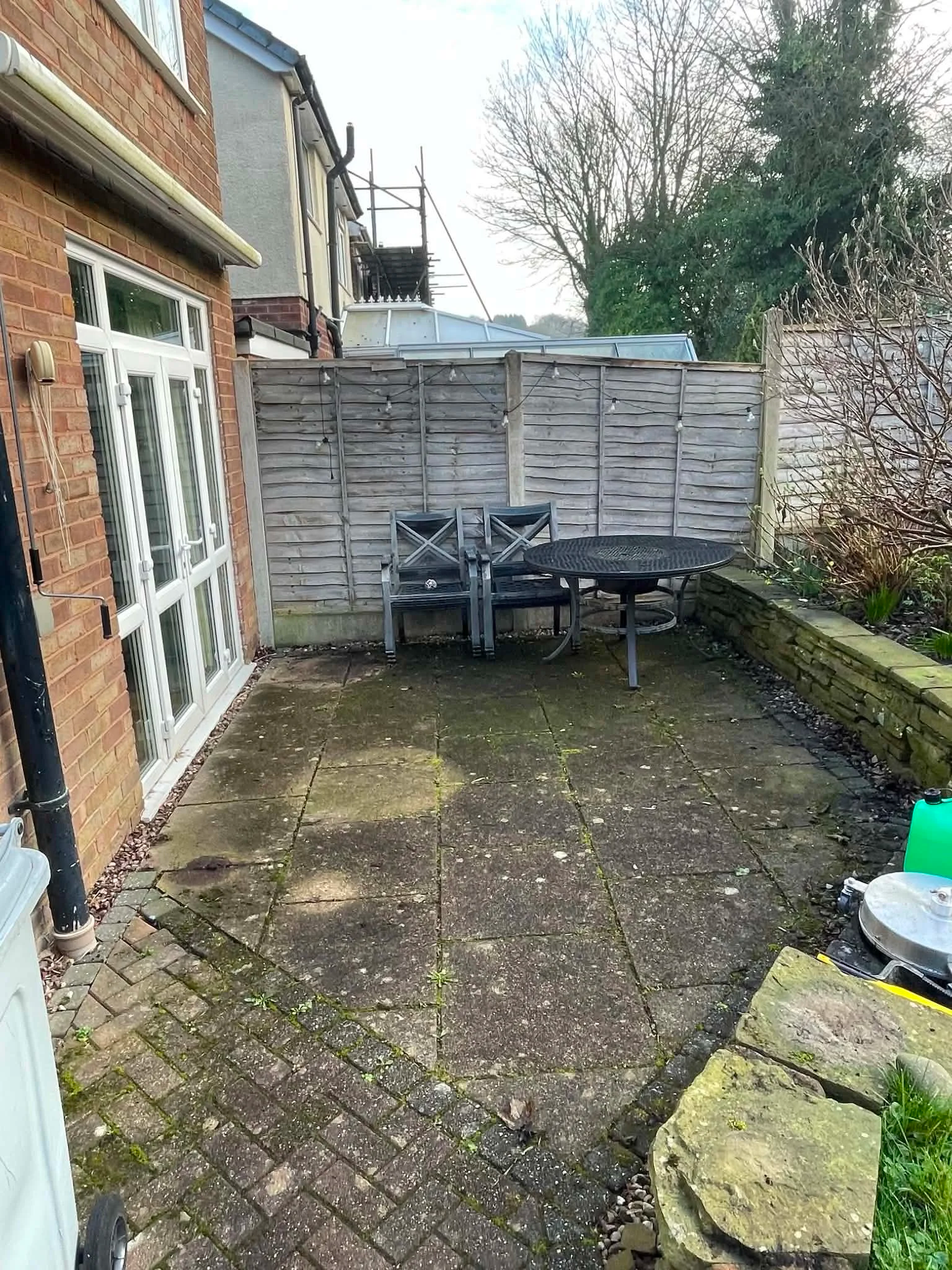 Backyard patio with outdoor furniture, including two chairs and a round table, surrounded by a wooden fence, a brick house wall with a sliding glass door, and garden plants and trees in the background.