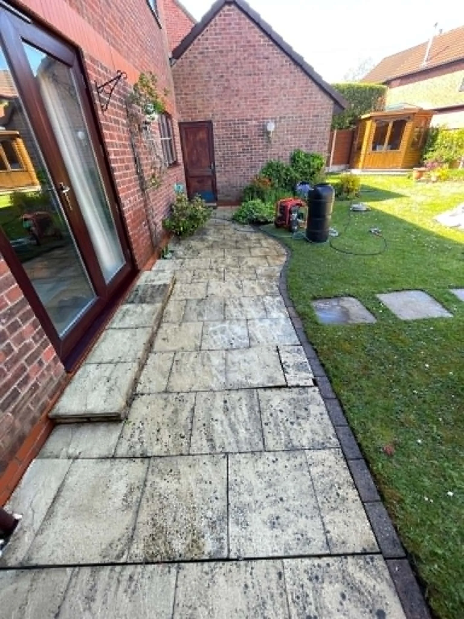 A backyard patio with a paved stone walkway, green lawn, and garden plants. The brick house wall has a glass door and decorative wall hanging. There is a wooden shed and various gardening tools and equipment.