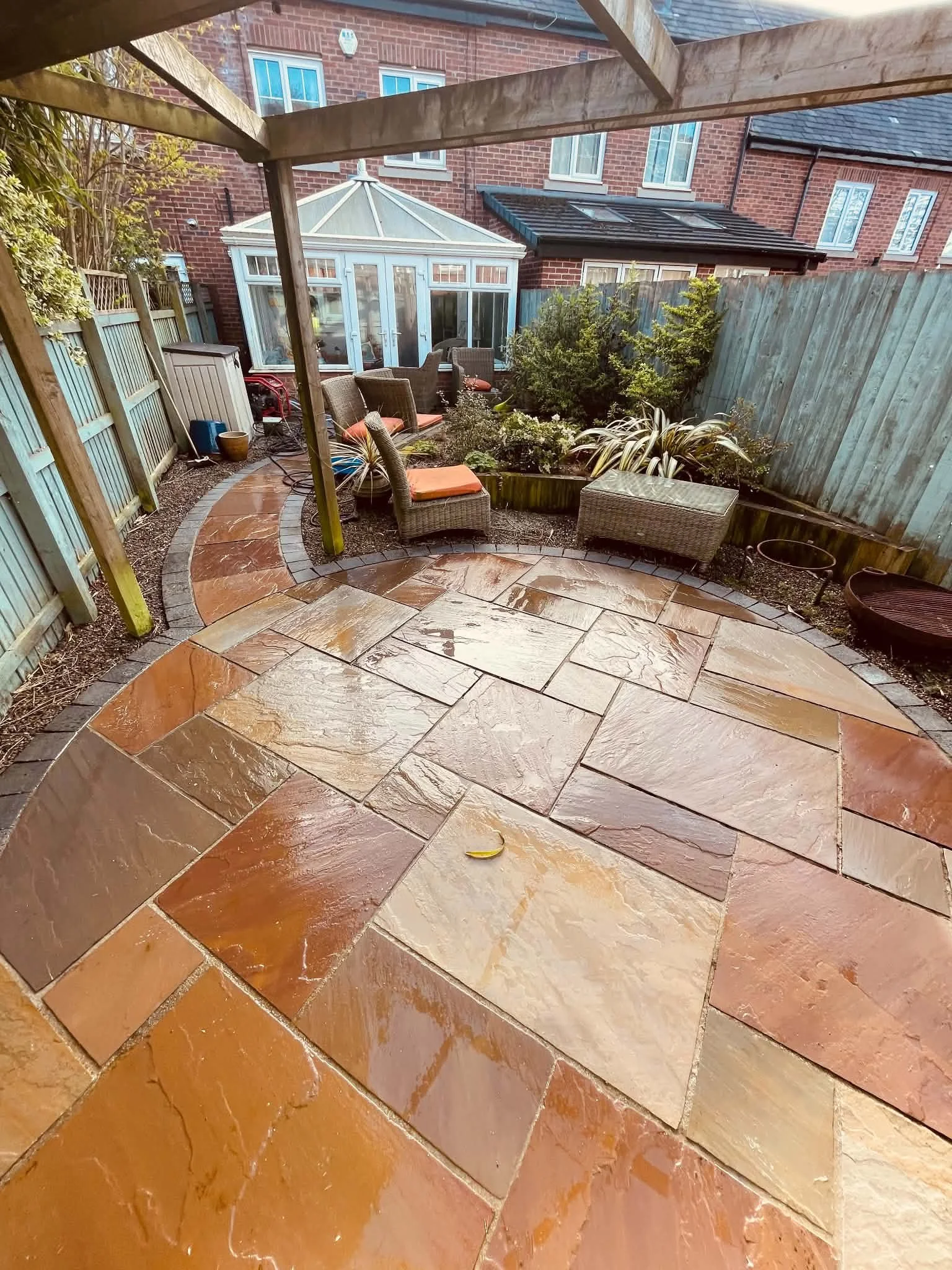 A backyard patio with curved stone paving, wicker chairs with orange cushions, a small garden bed with shrubs, and a wooden fence.