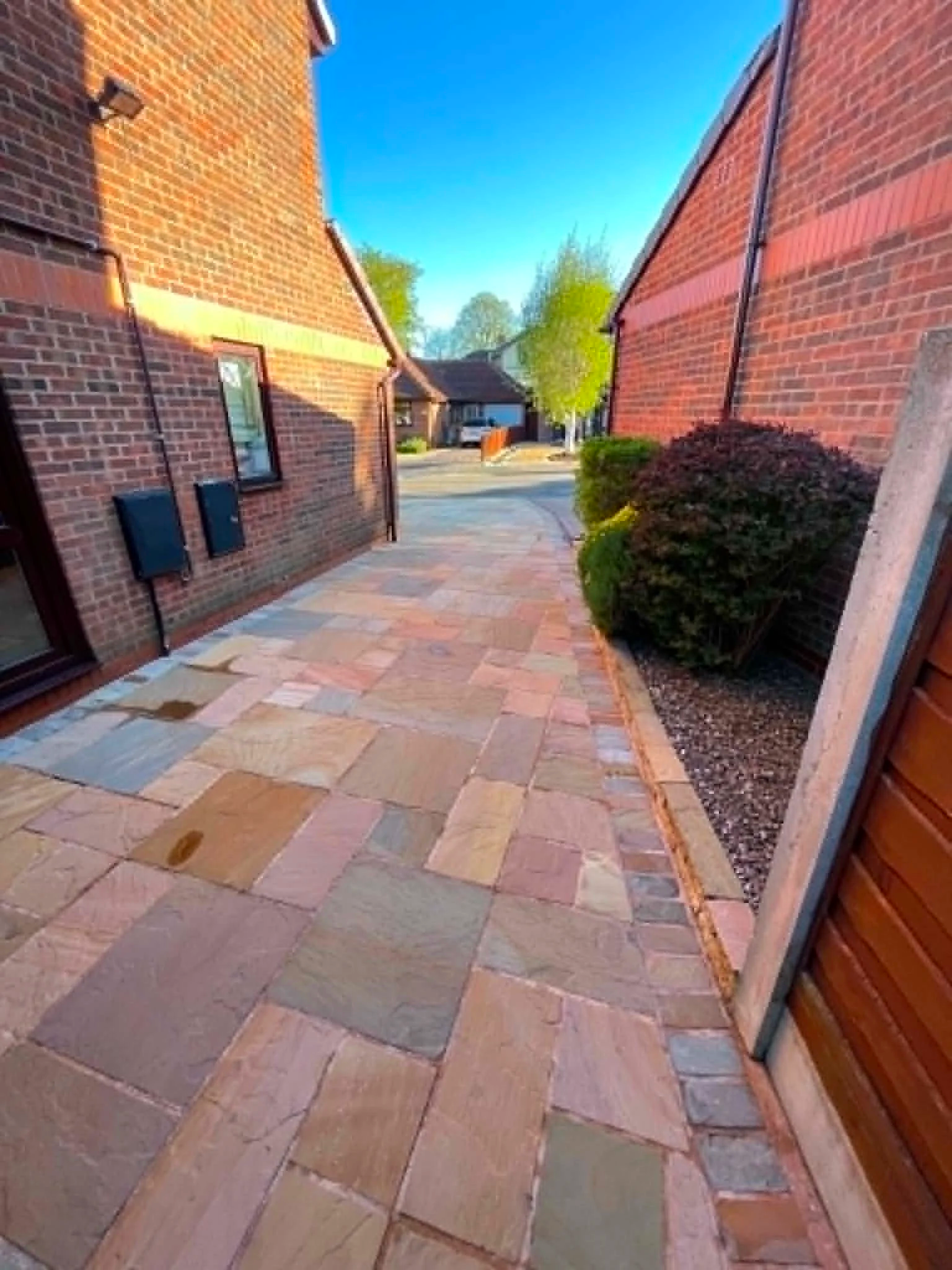 A view of a backyard patio area with multi-colored stone tiles, two brick walls on either side, bushes, and a house with trees in the background under a blue sky.