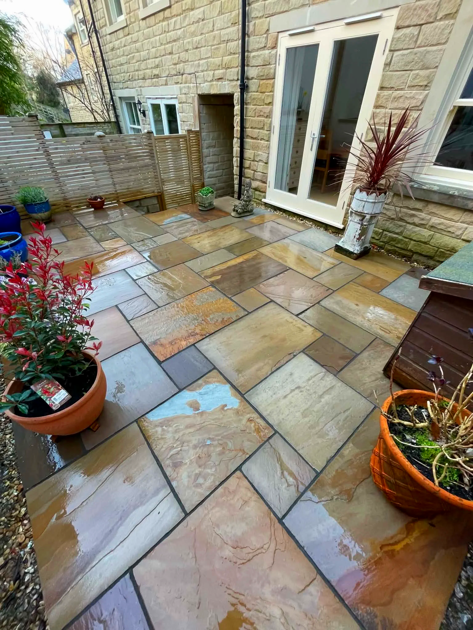 A wet stone patio with colorful tiles, potted plants, a small concrete Buddha statue, and a sliding glass door leading indoors.