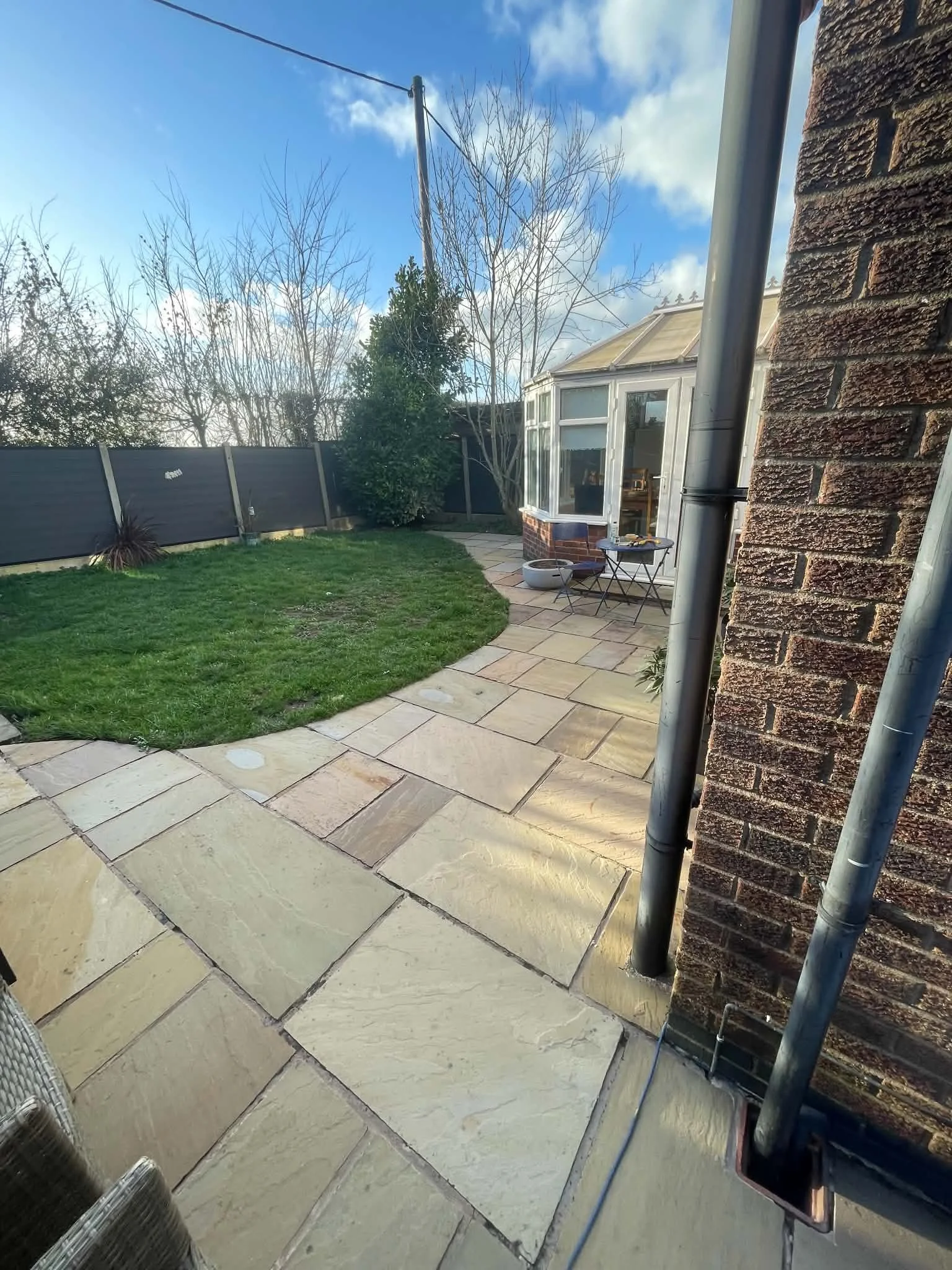 Backyard patio with beige stone tiles, green grass, a black fence, small trees, and a white conservatory with large windows. Sky is partly cloudy with some blue visible.