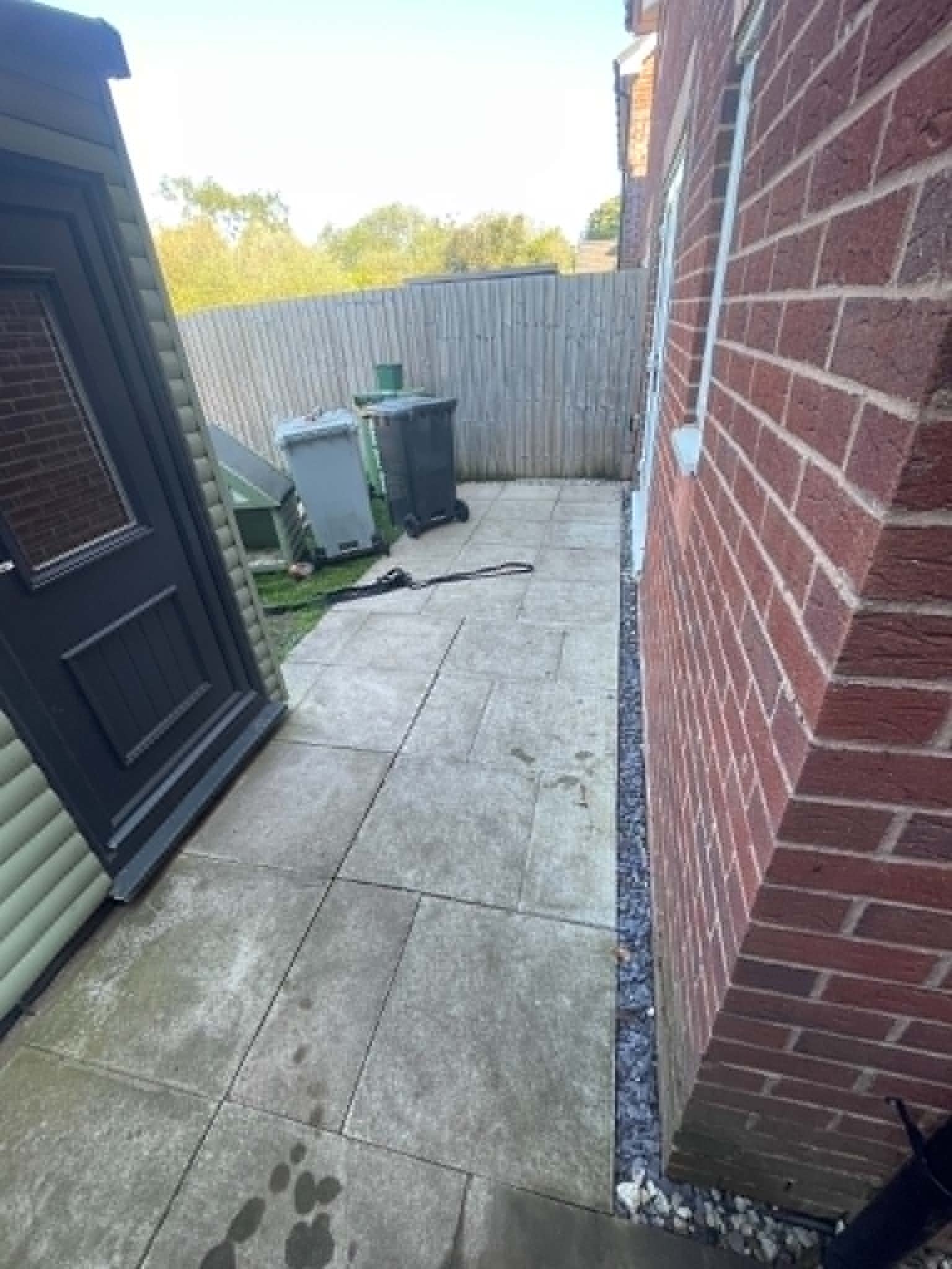 A small outdoor patio area with a brick wall on the right and green siding on the left, tiled with large concrete slabs, and a metal fence at the back. Two garbage bins, one gray and one black, are near the fence. A green watering can is on top of the gray bin. Dark spots on the ground appear to be water or oil stains.