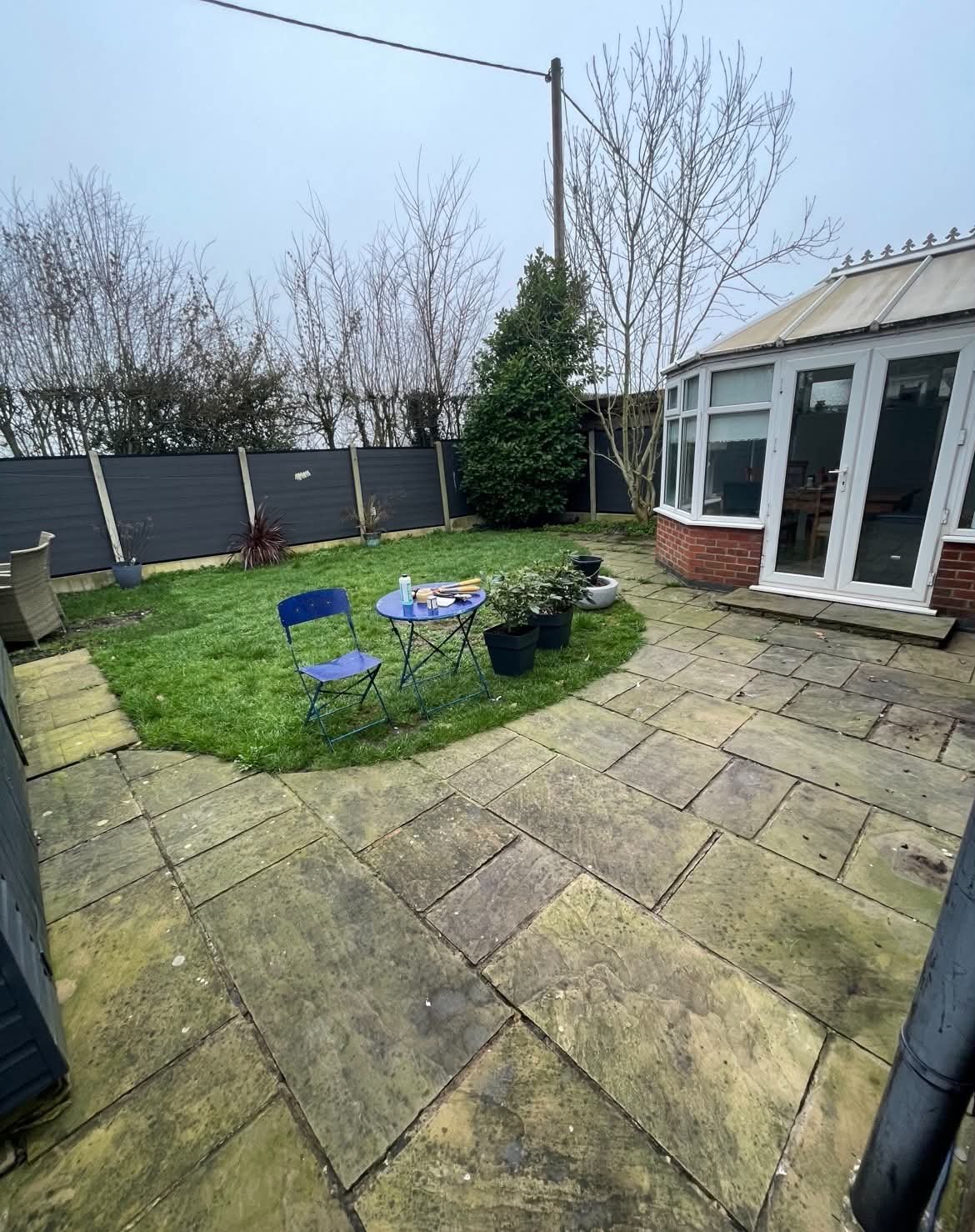 A backyard patio with stone paving, a small grass patch, outdoor furniture, potted plants, and a conservatory attached to a brick house, surrounded by a black fence and leafless trees in the background.