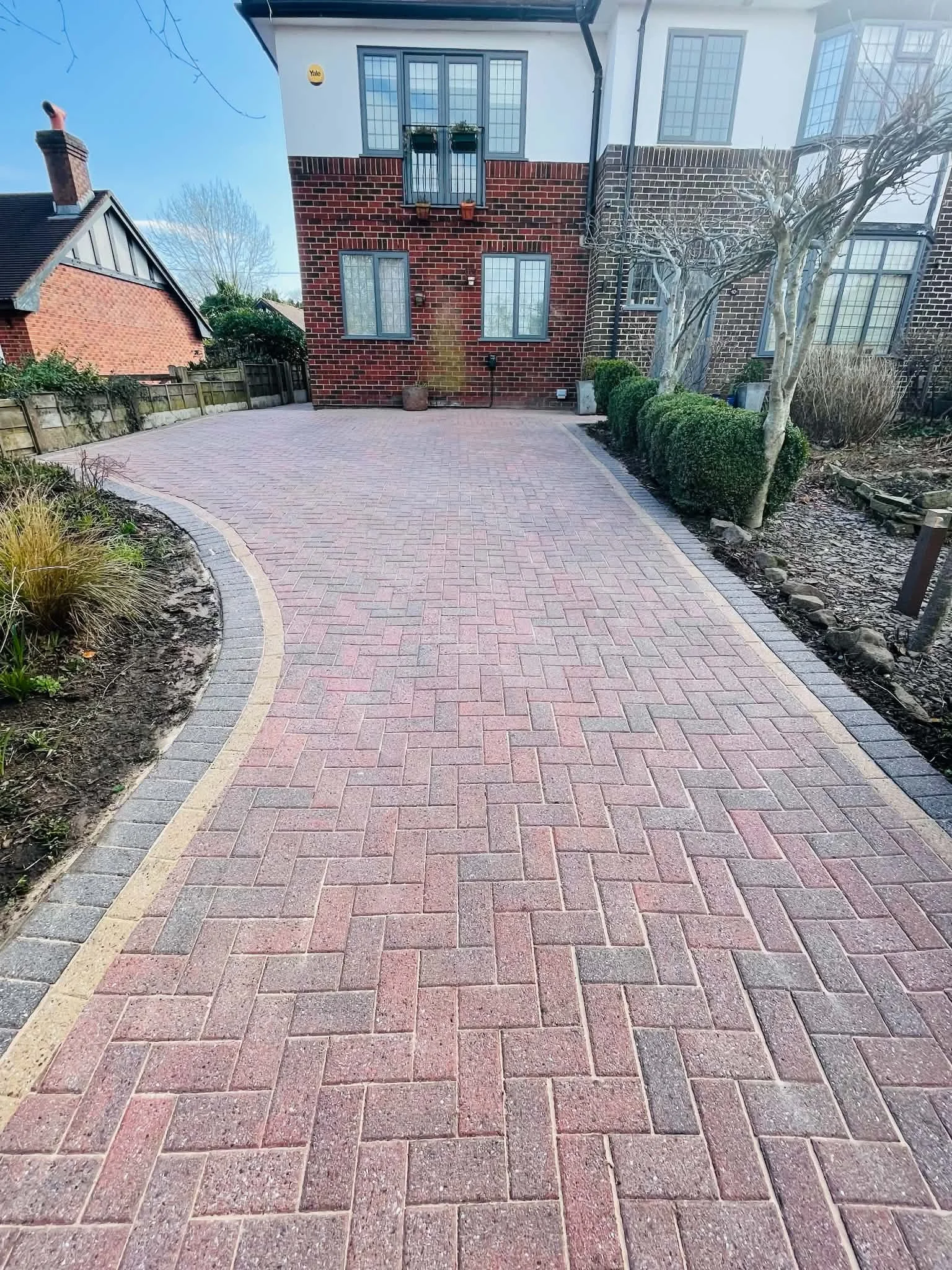 A paved brick driveway with a curved border, leading to the back of a house with brick walls and multiple windows. There are small trees and bushes to the right of the driveway and a small section of garden on the left. The house has a white upper part and a brick lower part, with some potted plants and garden decorations visible.