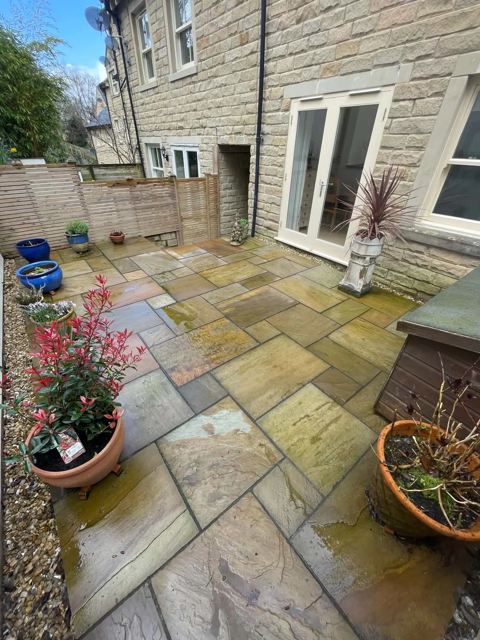 A small outdoor patio area with wet sandstone tiles, potted plants, a wooden fence, a stone wall house with white-framed windows and a glass door, garden decorative objects, and greenery around.