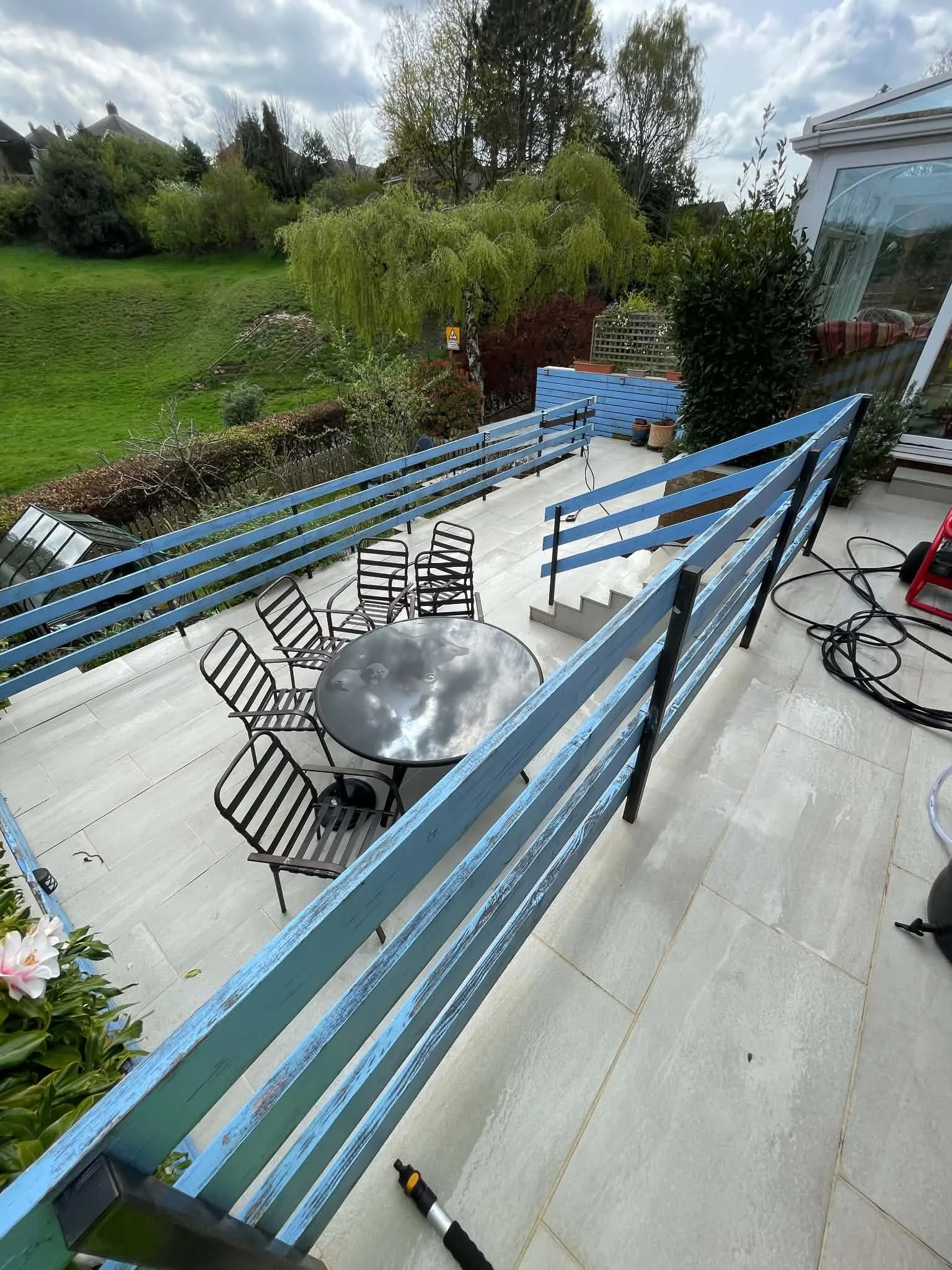 View of an outdoor balcony with a round table and several black chairs, a staircase, potted plants, a blue railing, and a green backyard with trees and grass in the background.