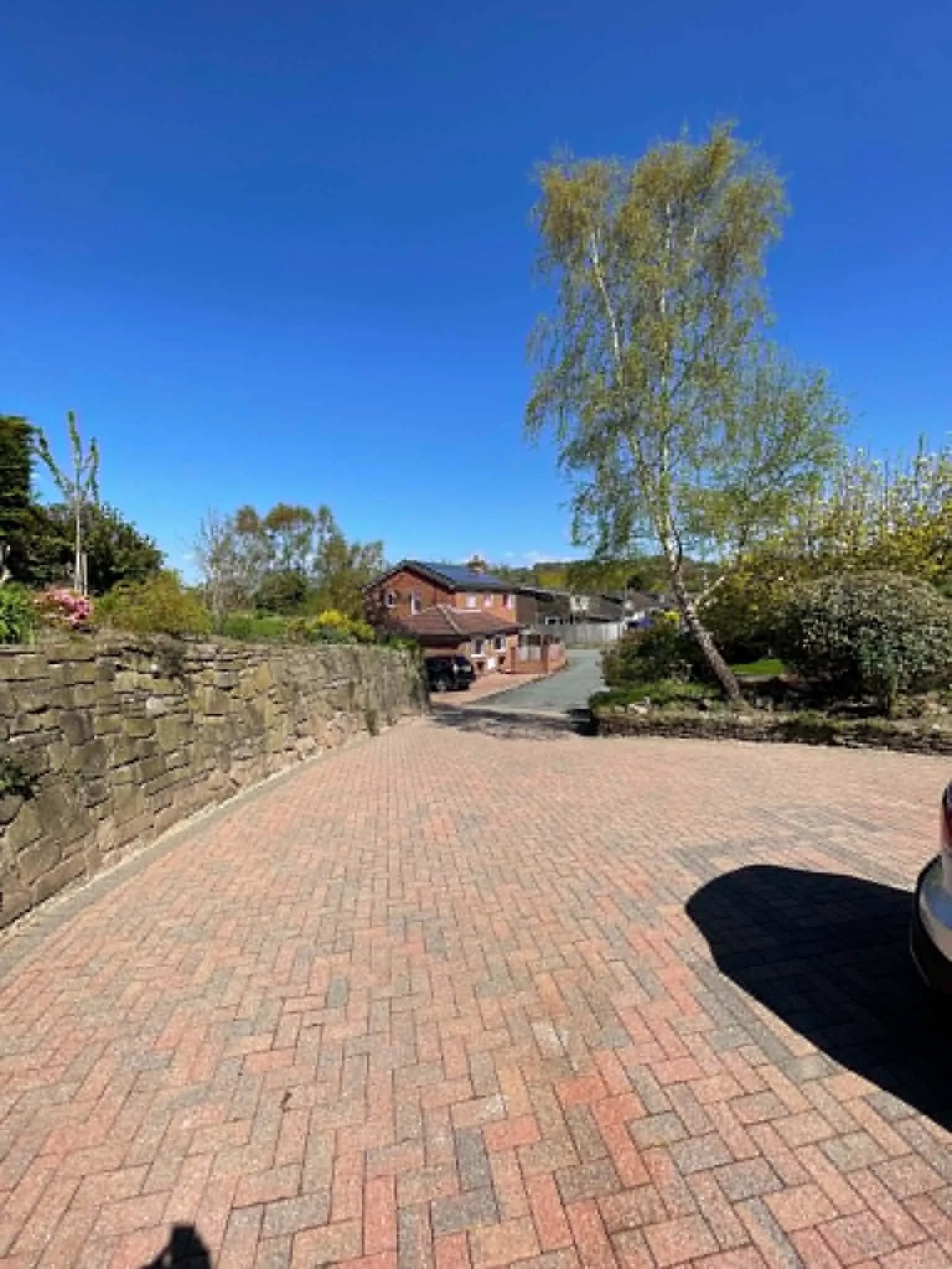 A paved brick driveway with stone walls on the sides, leading to a house in the background with a sloped roof, surrounded by trees and bushes under a clear blue sky.
