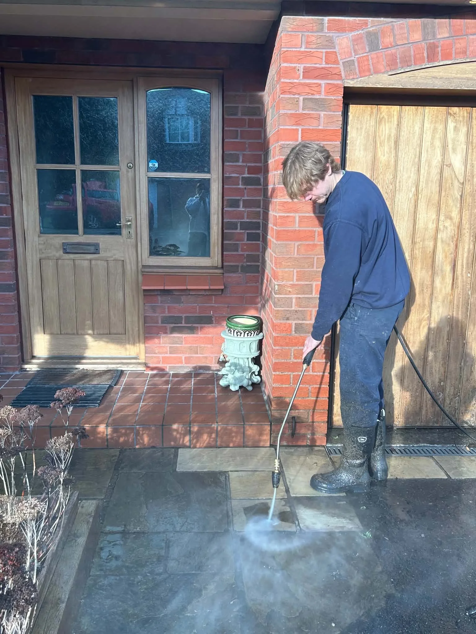 A person cleaning a concrete patio with a pressure washer, standing on the patio in front of a brick and wooden house exterior.
