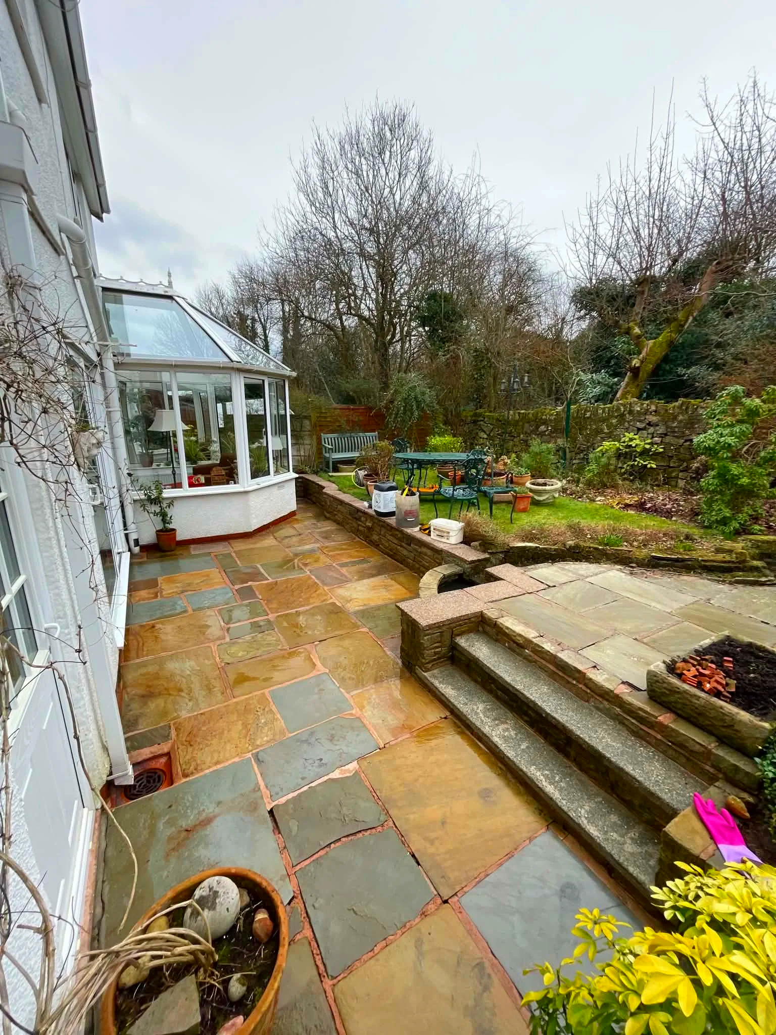 A view of a backyard patio with multicolored stone tiles, potted plants, gardening tools, a picnic table, and bare trees in the background under an overcast sky.