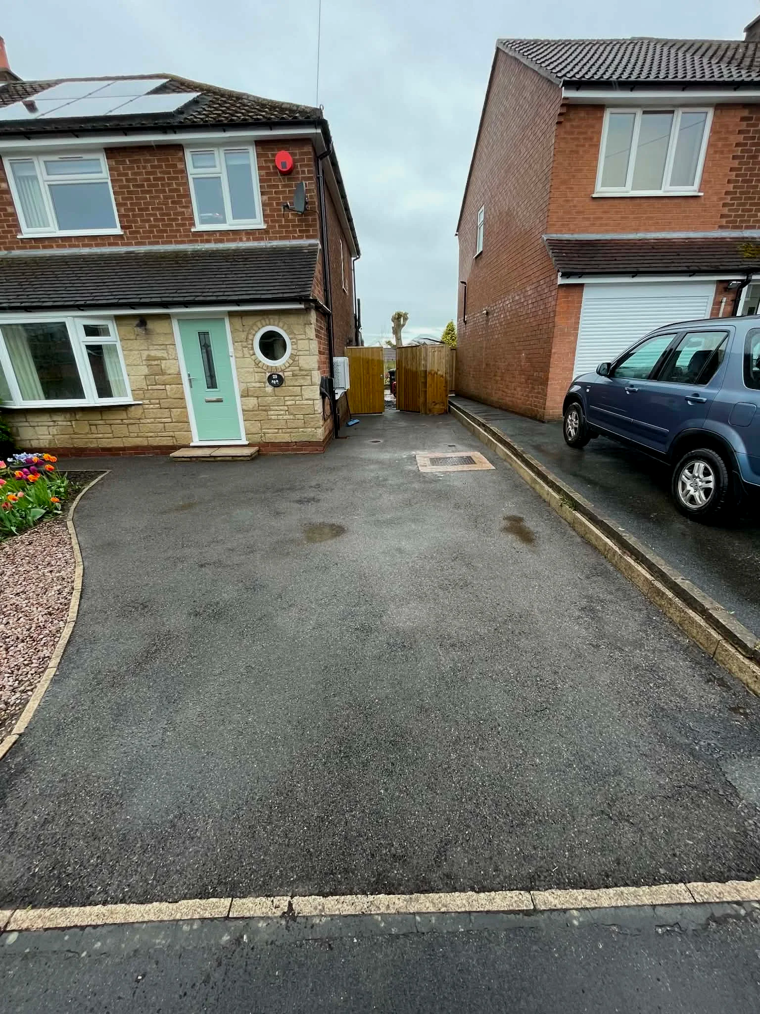 View of a driveway with a parked blue car next to brick houses, a flower bed with tulips, and a wooden gate at the back.