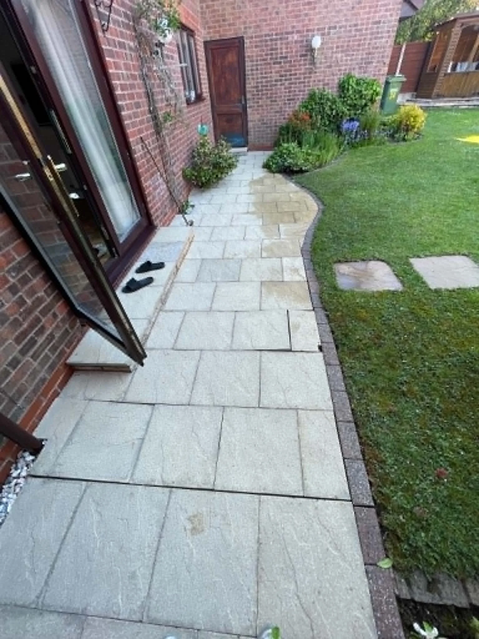 A small paved patio area next to a brick house wall with a window, a door, some potted plants, and a grassy lawn with stepping stones.