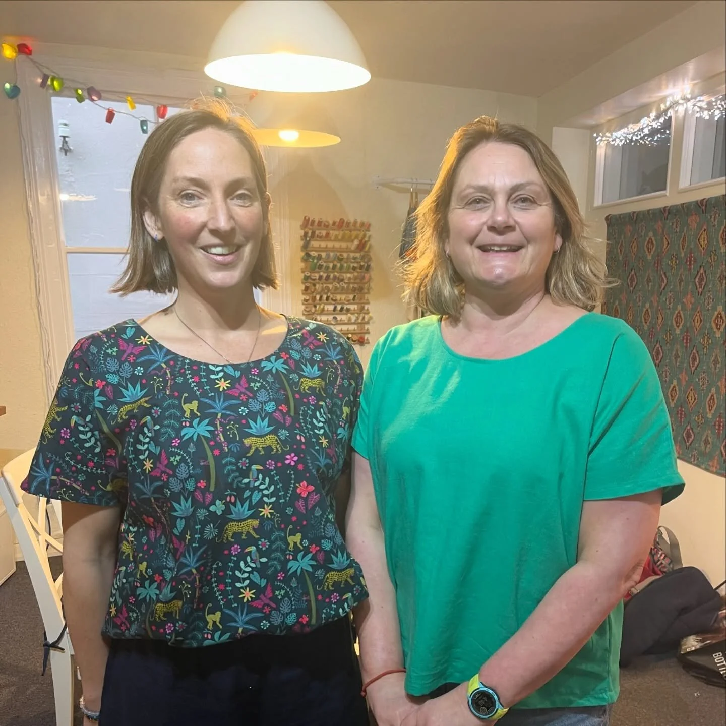 Two women smiling in a room decorated with string lights and a window with colorful garland.