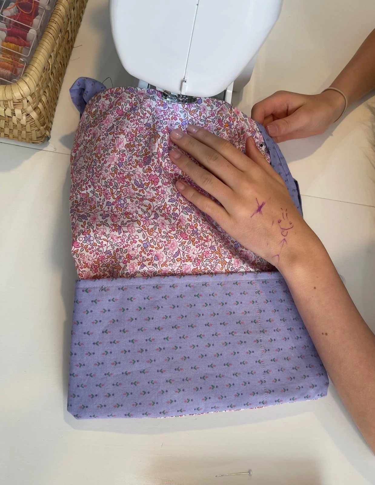 A person sewing fabric with a sewing machine on a white table. The fabric has a floral pattern with pink, purple, and orange colors, and the person's hand is resting on the fabric. There is a basket with sewing supplies nearby.