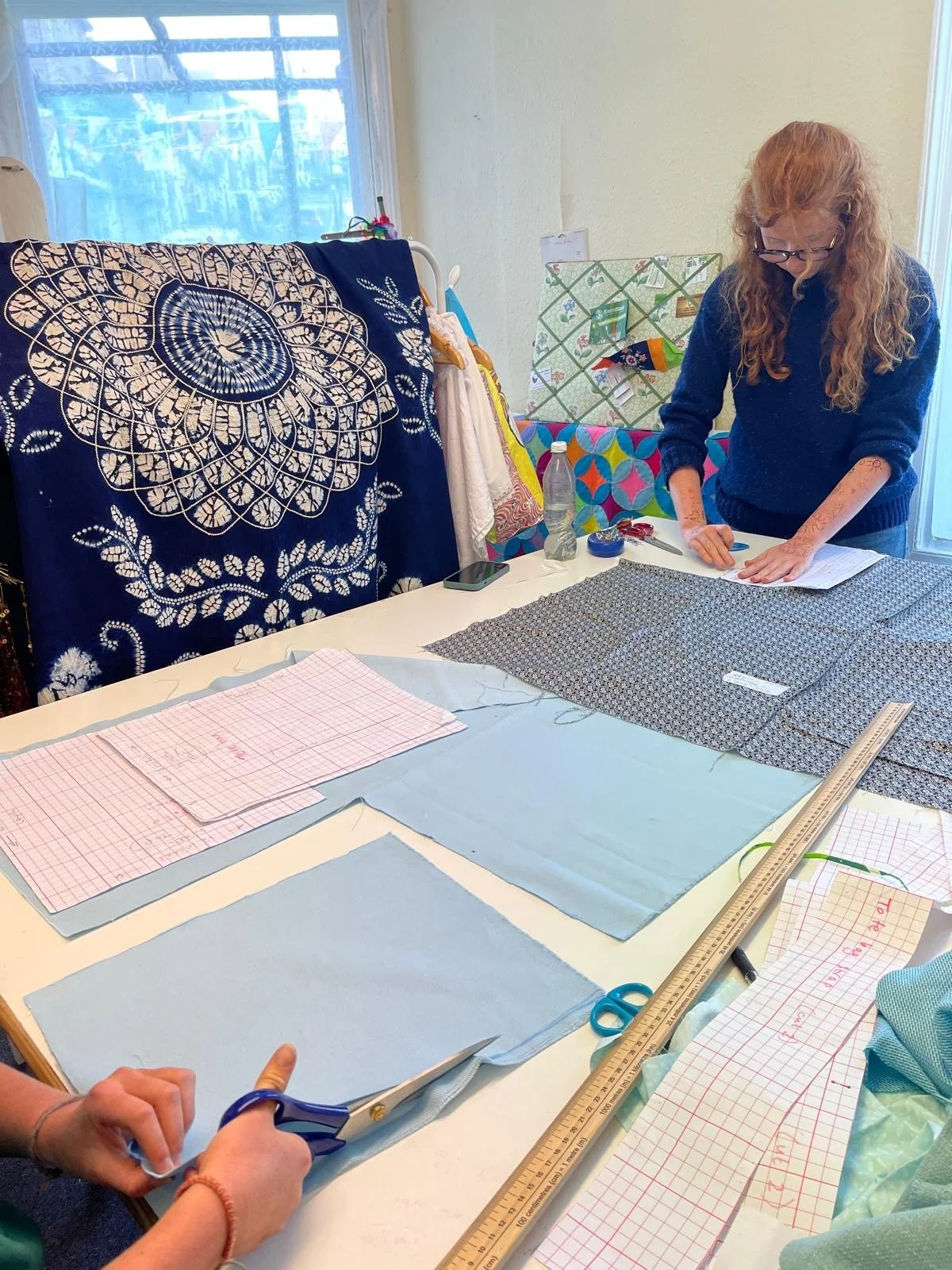 A woman with red hair, wearing glasses and a blue sweater, working at a table with fabric, patterns, scissors, a ruler, and paper for sewing or crafting.