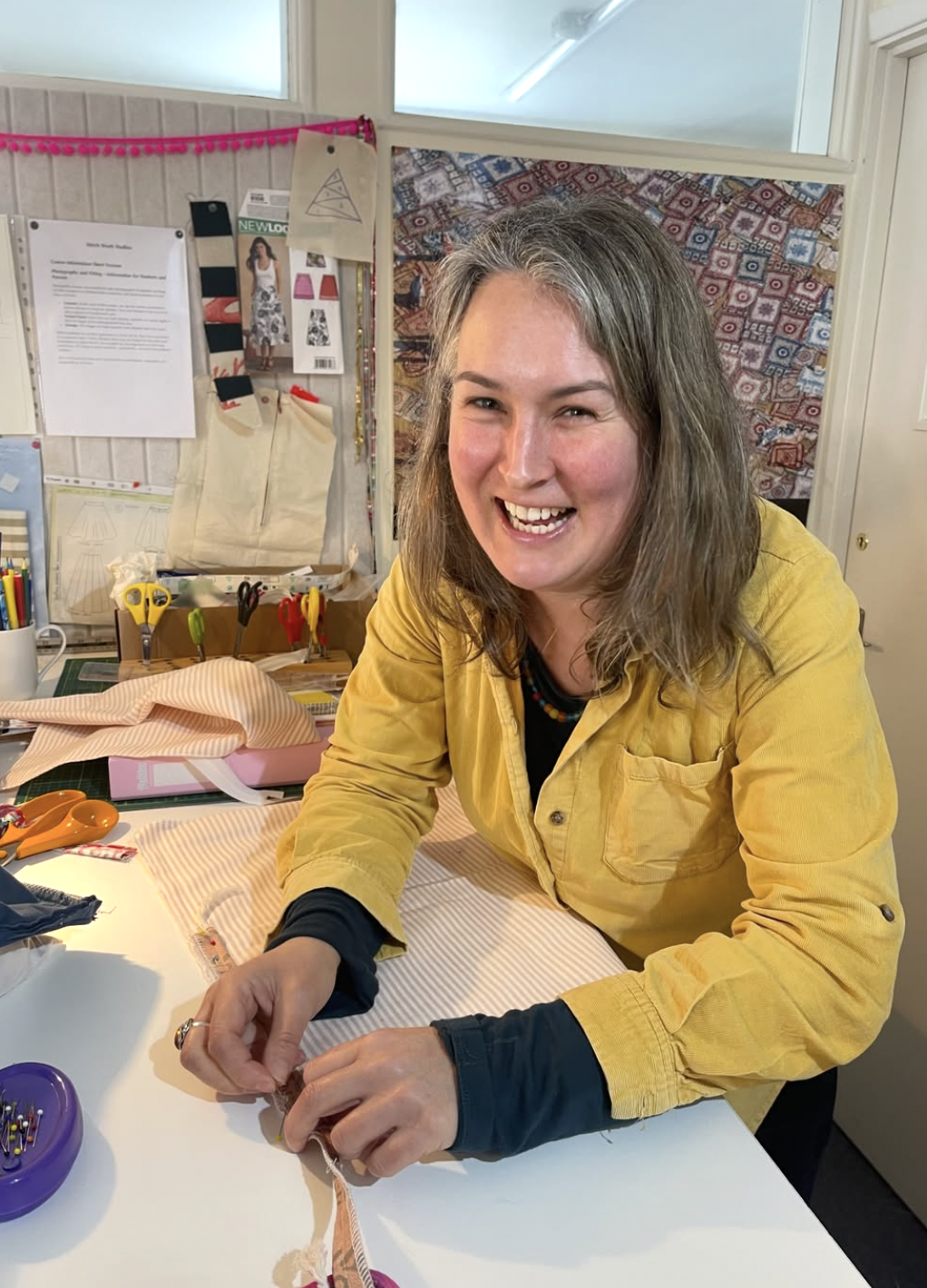 Smiling woman sewing fabric at a crafting table, surrounded by sewing supplies and fabric patterns.