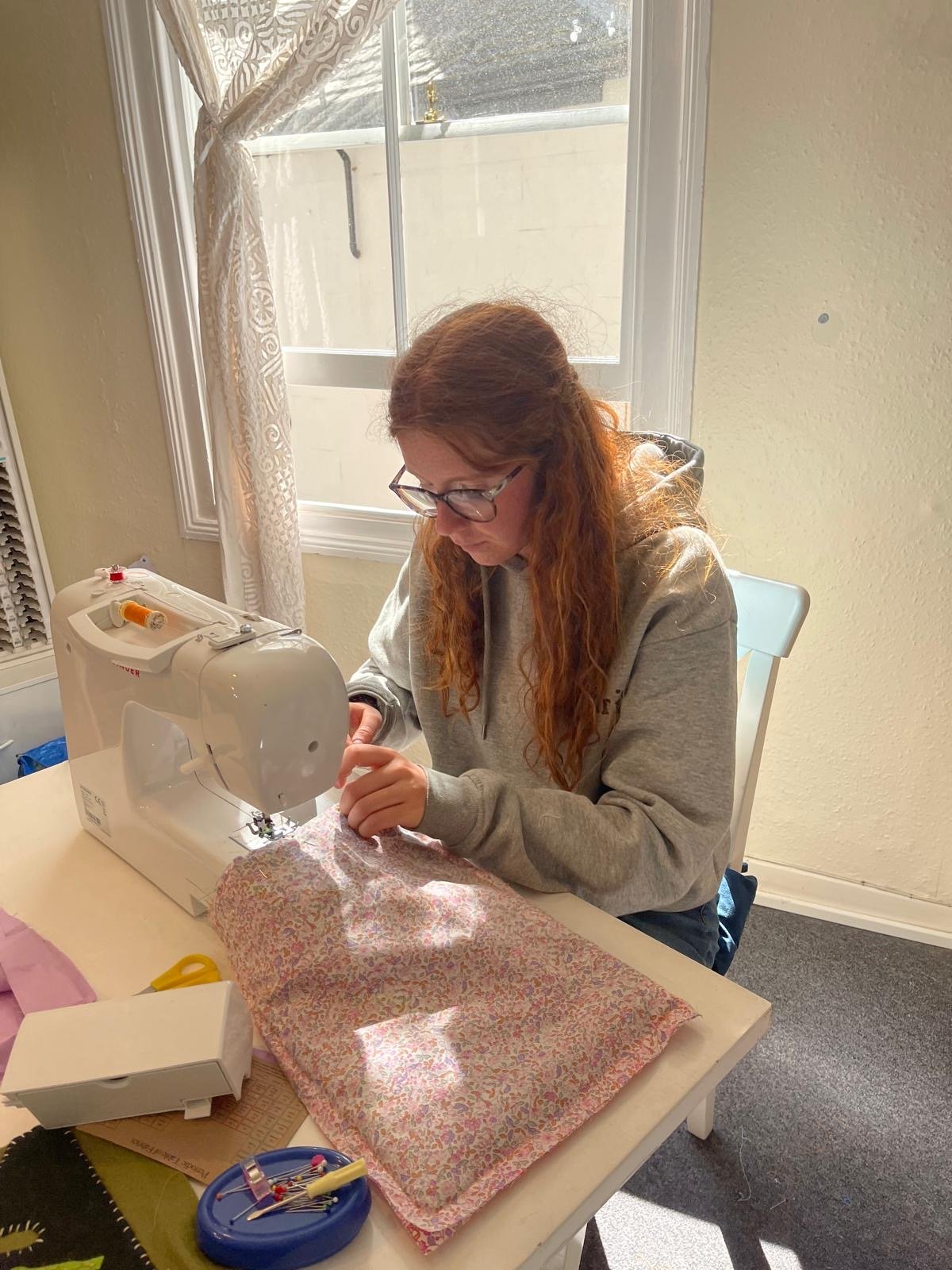 A woman sewing with a sewing machine at a table, wearing glasses and a grey hoodie, in a room with a window and beige walls.
